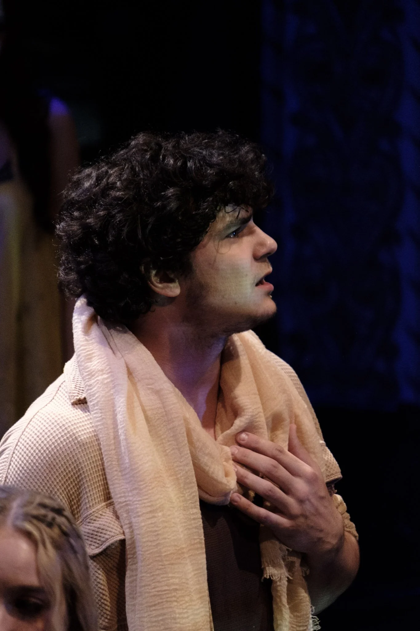 A man with curly dark hair has his hand on his chest and appears emotional, possibly crying, in a scene from a theatrical play.