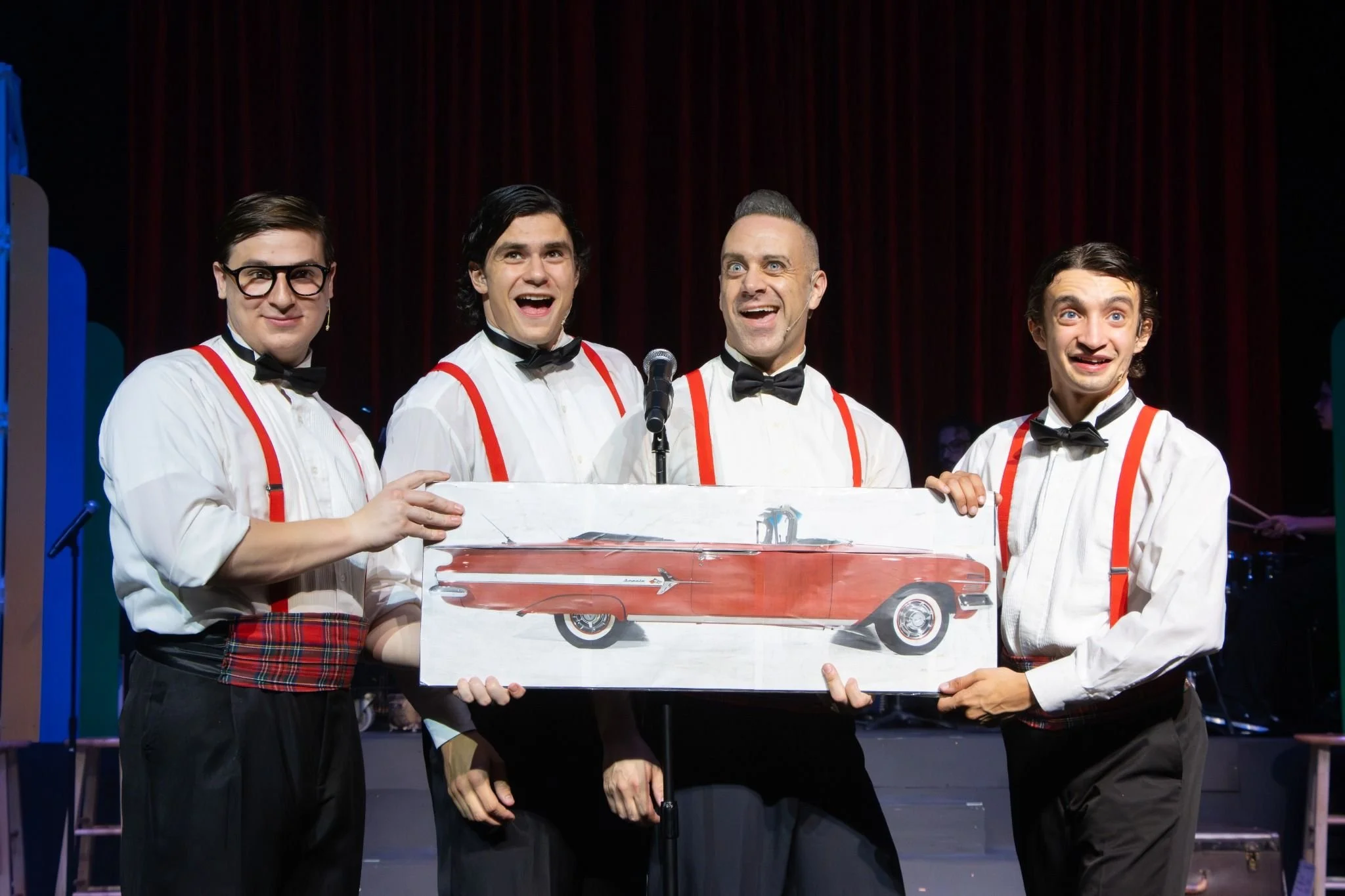 Four men dressed in white shirts, black bow ties, red suspenders, and plaid kilts on stage, holding a sign with a red vintage car illustration, with a red curtain backdrop.