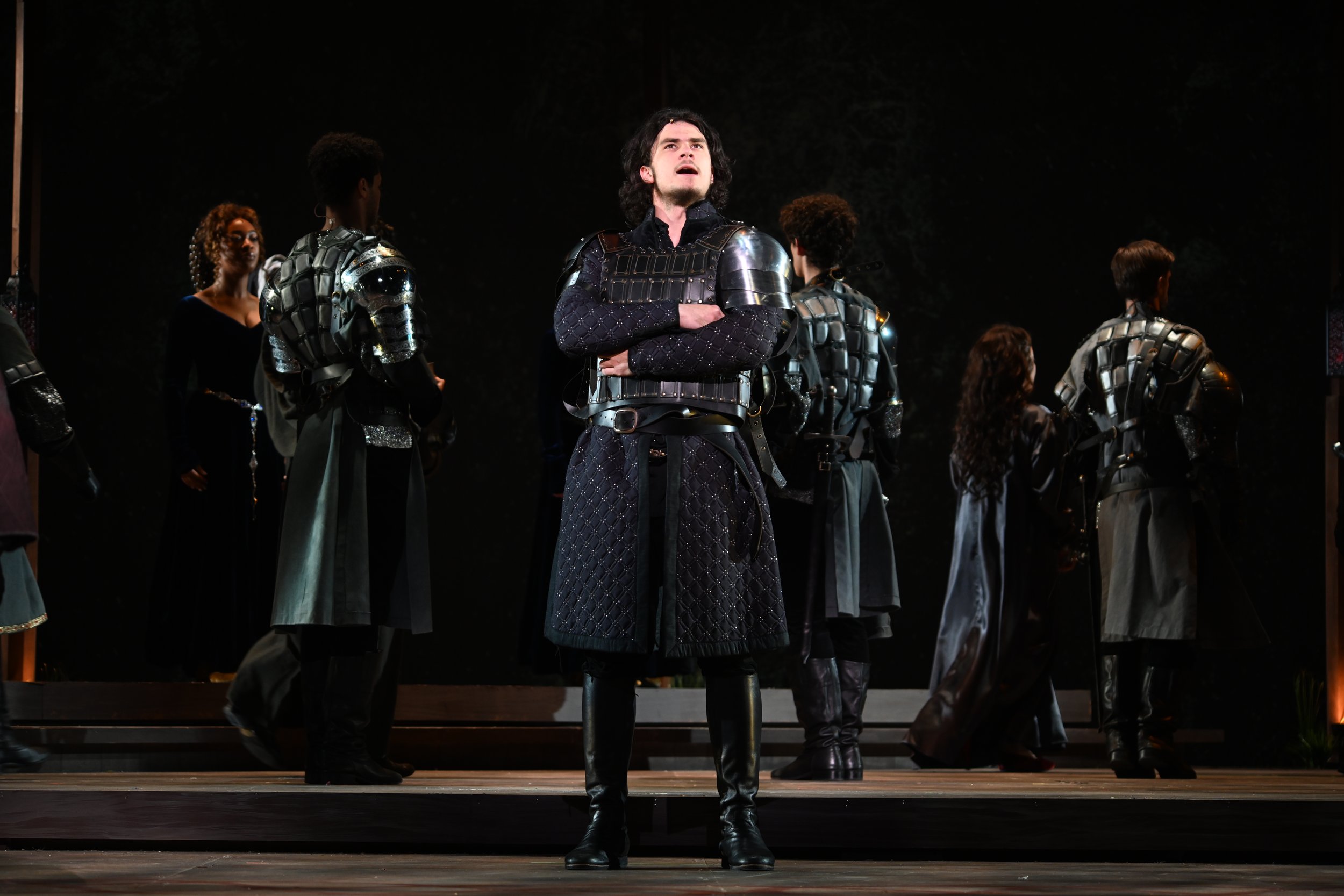 A man standing on stage with his arms crossed over his chest, surrounded by people in dark, medieval-style costumes with armor.