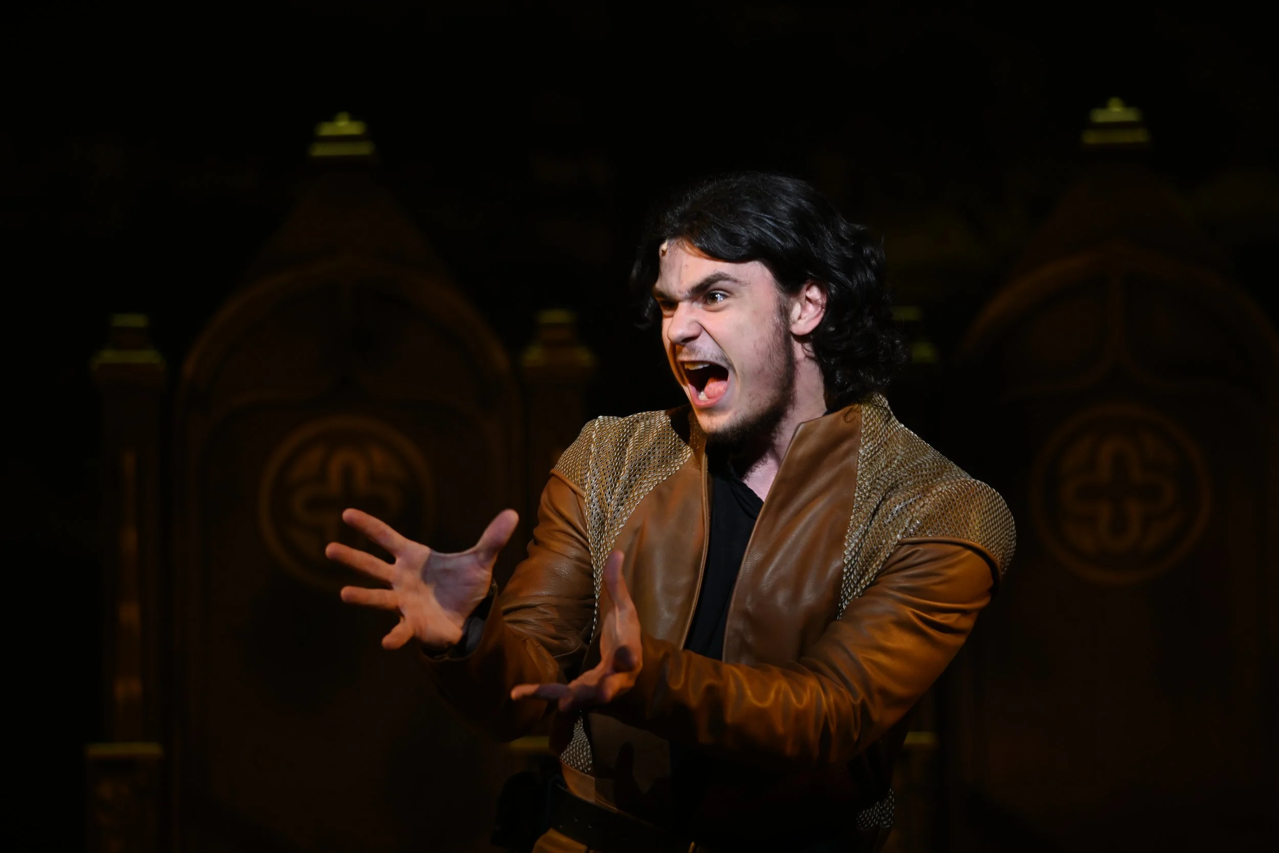 A man with black hair and a beard, wearing a brown leather jacket, appears to be shouting or yelling with an angry or intense expression. He is on a stage with a dark, ornate background.
