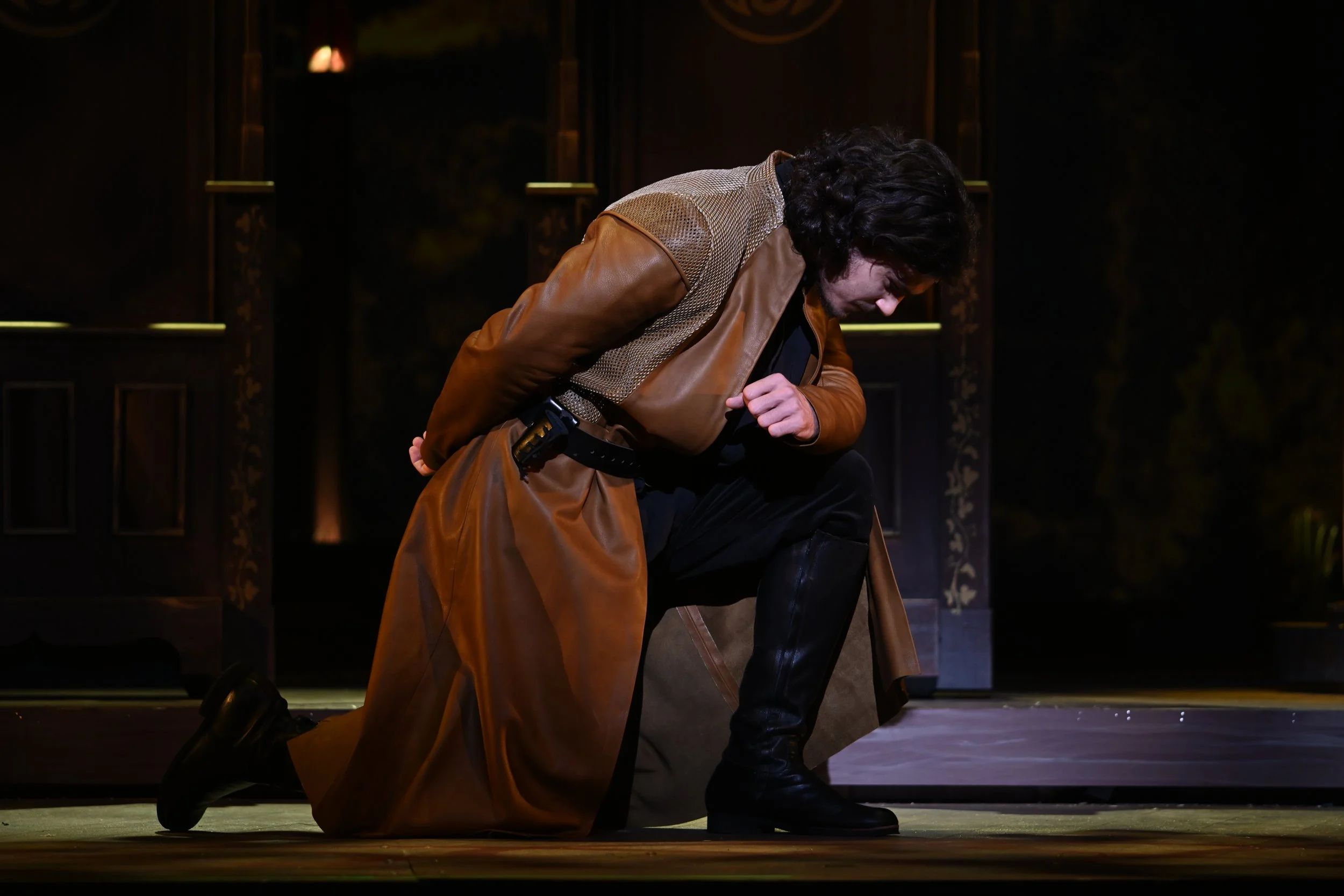 A man dressed in a brown leather coat and black boots is kneeling on stage, appearing to be in emotional distress, with his head bowed and hands clutching his coat.
