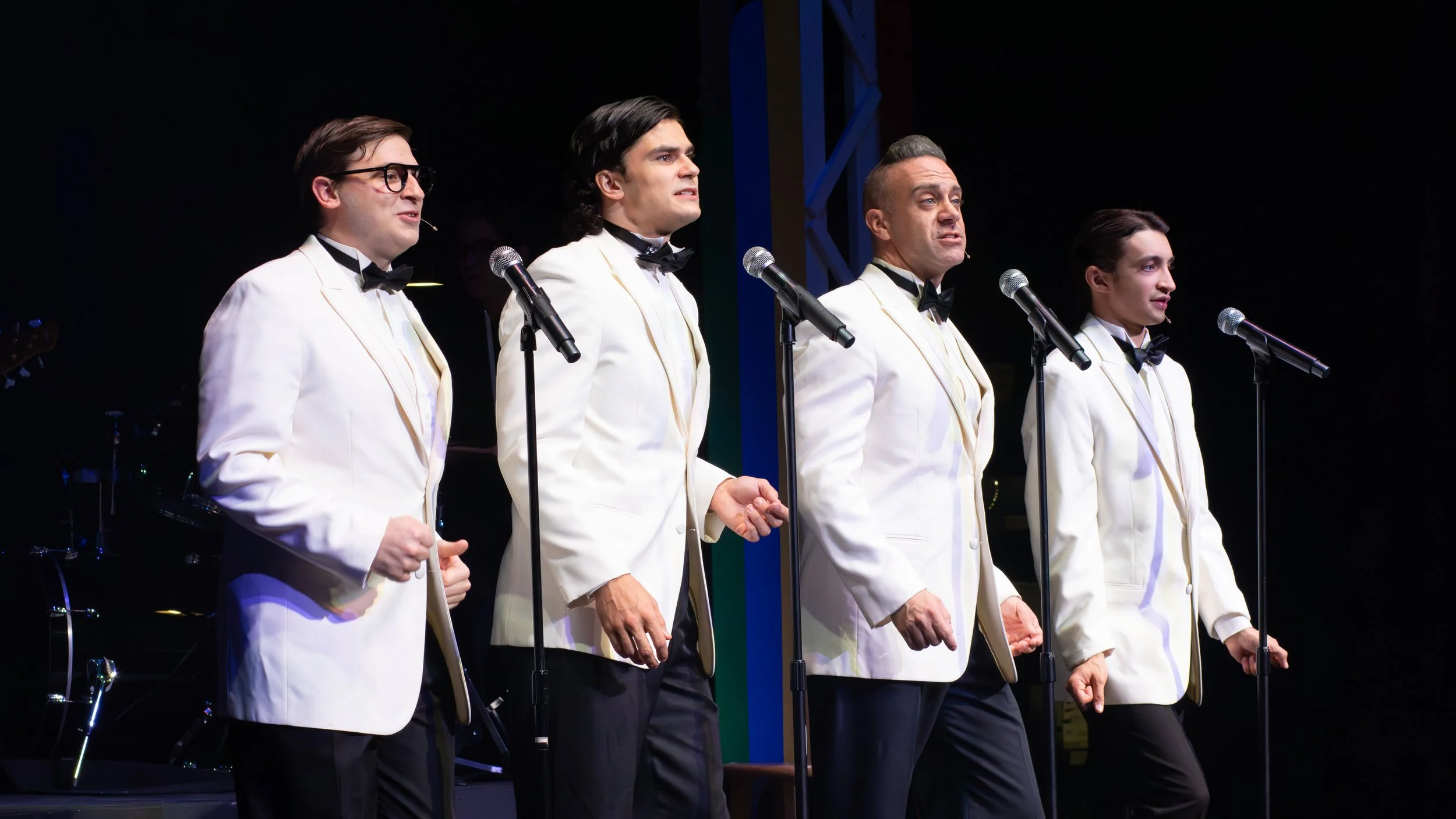 Four male performers dressed in white tuxedos with black bow ties, standing on stage with microphones, performing in front of a dark background.