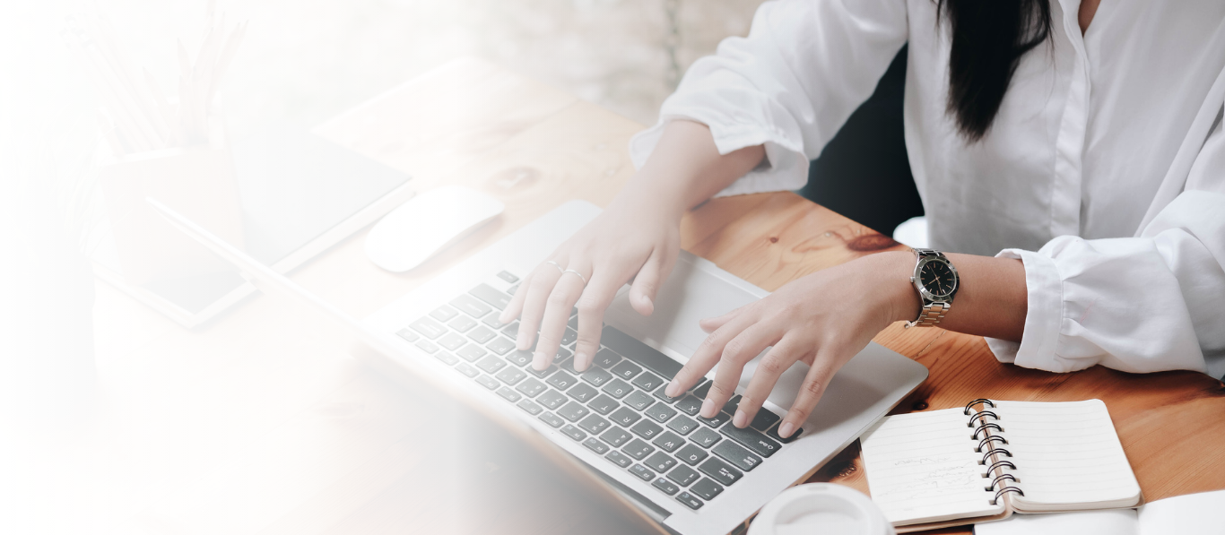 Person typing on a laptop at a wooden desk with a notebook, coffee cup, and other items.