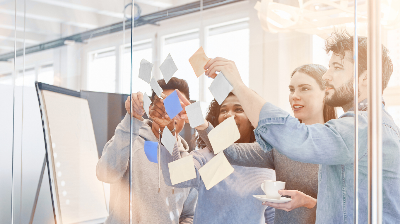 A diverse group of five young people working together on a project in a modern office, sticking notes on a glass wall.