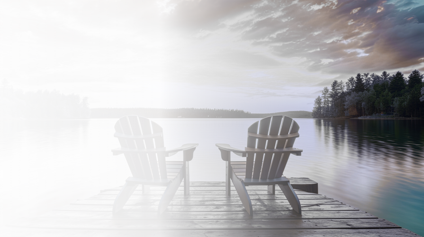 Two Adirondack chairs facing a calm lake with a dock, surrounded by trees under a partly cloudy sky, during sunset or sunrise.