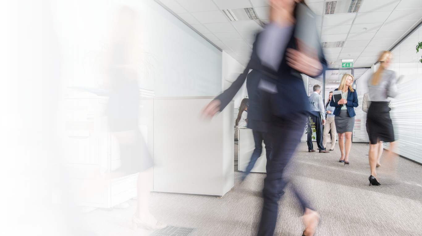 Business professionals walking and standing in a modern office corridor, some holding documents and others engaged in conversation.