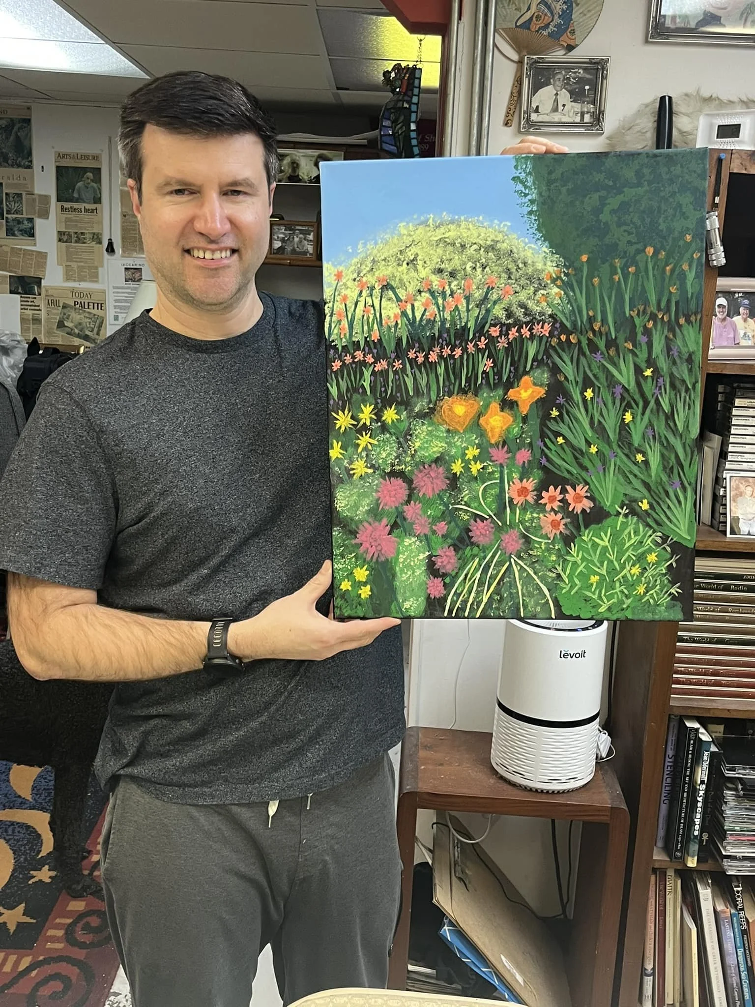 A student of Raphael Iaccarino's holding a colorful painting of a lush garden with flowers, bushes, trees, and a bright blue sky.