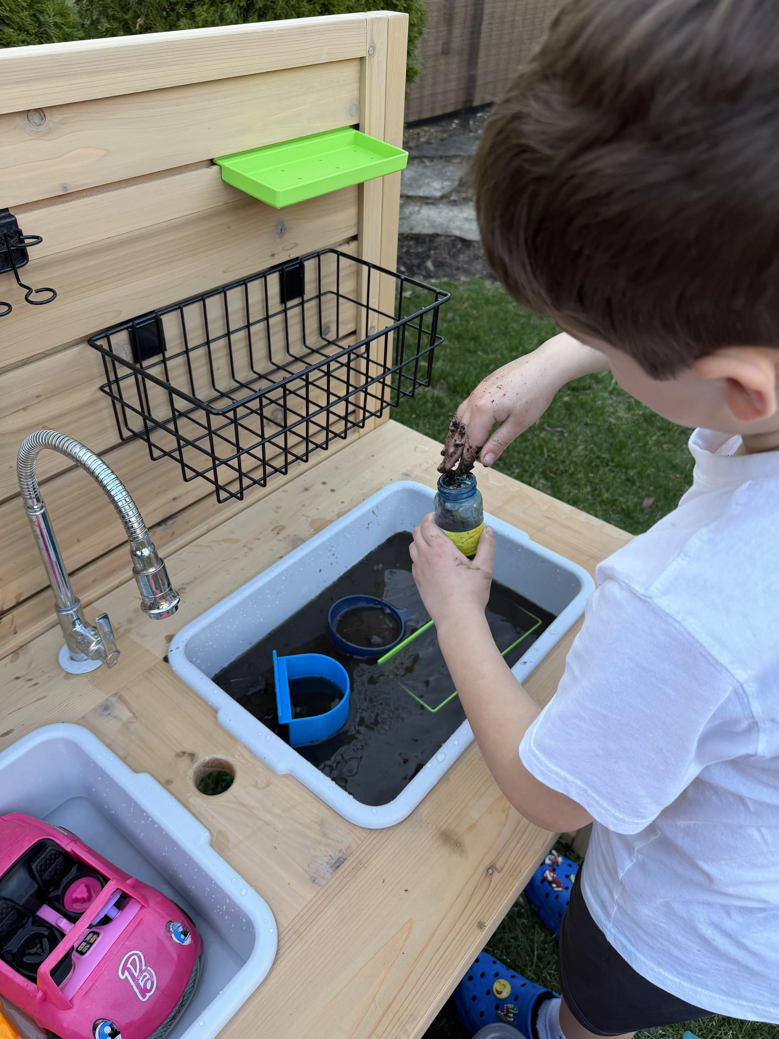 A Kinderbuilt cedar mud kitchen featuring a vertical activity slat wall with storage bins and a working water faucet.