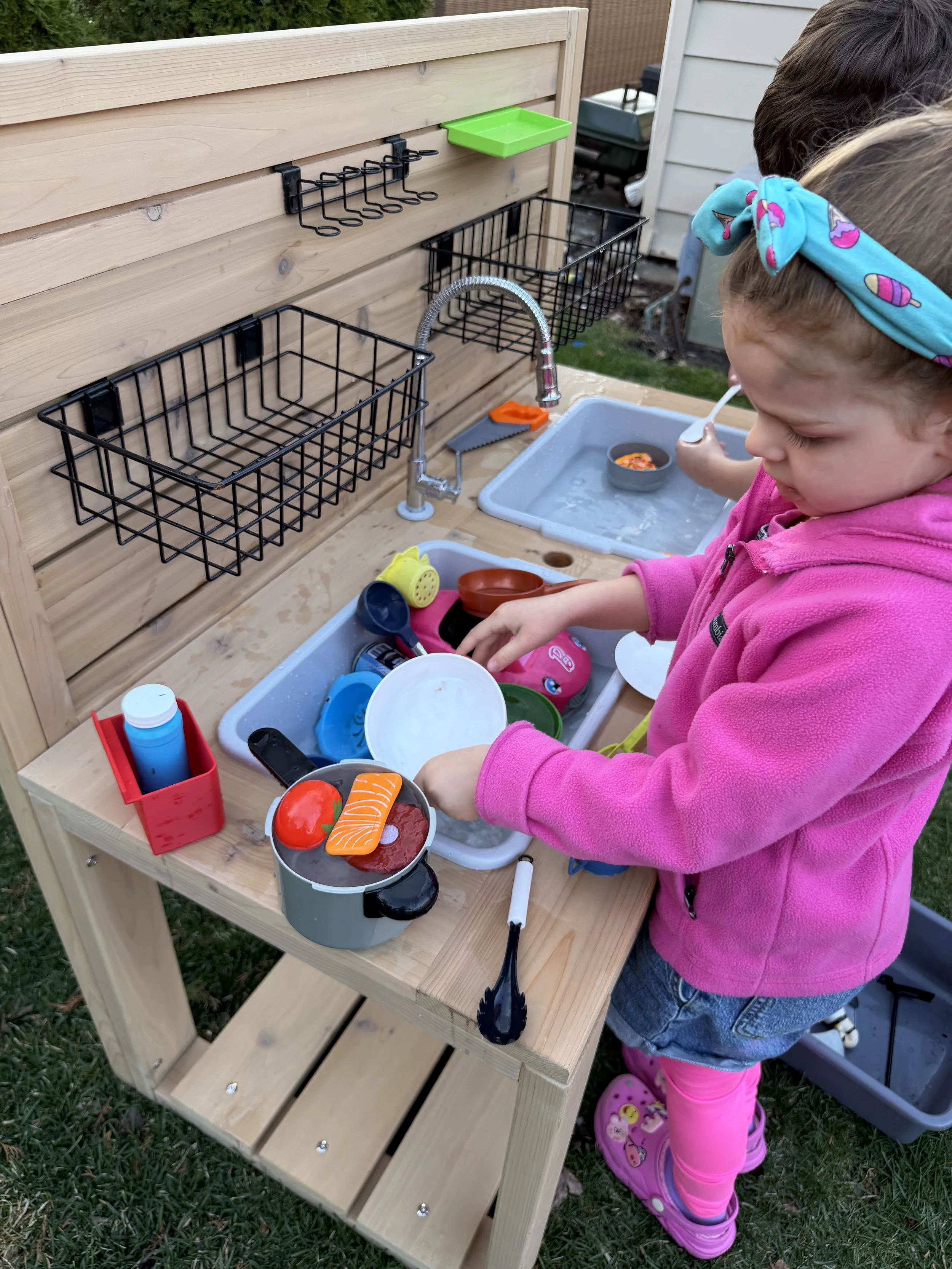 A young girl in a pink hoodie and pink socks playing at a wooden outdoor play kitchen, washing toy dishes and pretending to cook with plastic food.