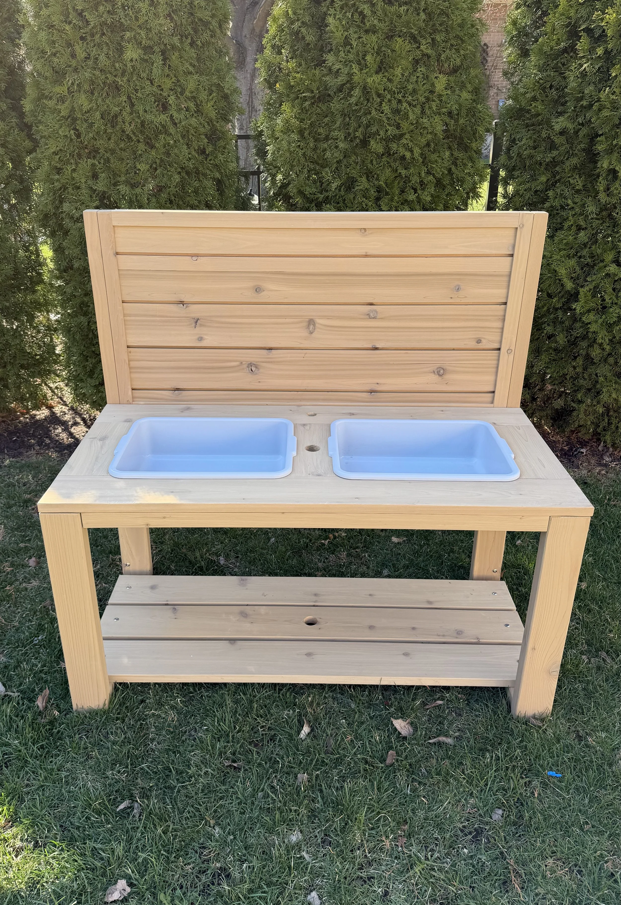 A wooden outdoor play kitchen with two blue plastic sinks, a backsplash, and an open shelf below on a grassy area, surrounded by green bushes.
