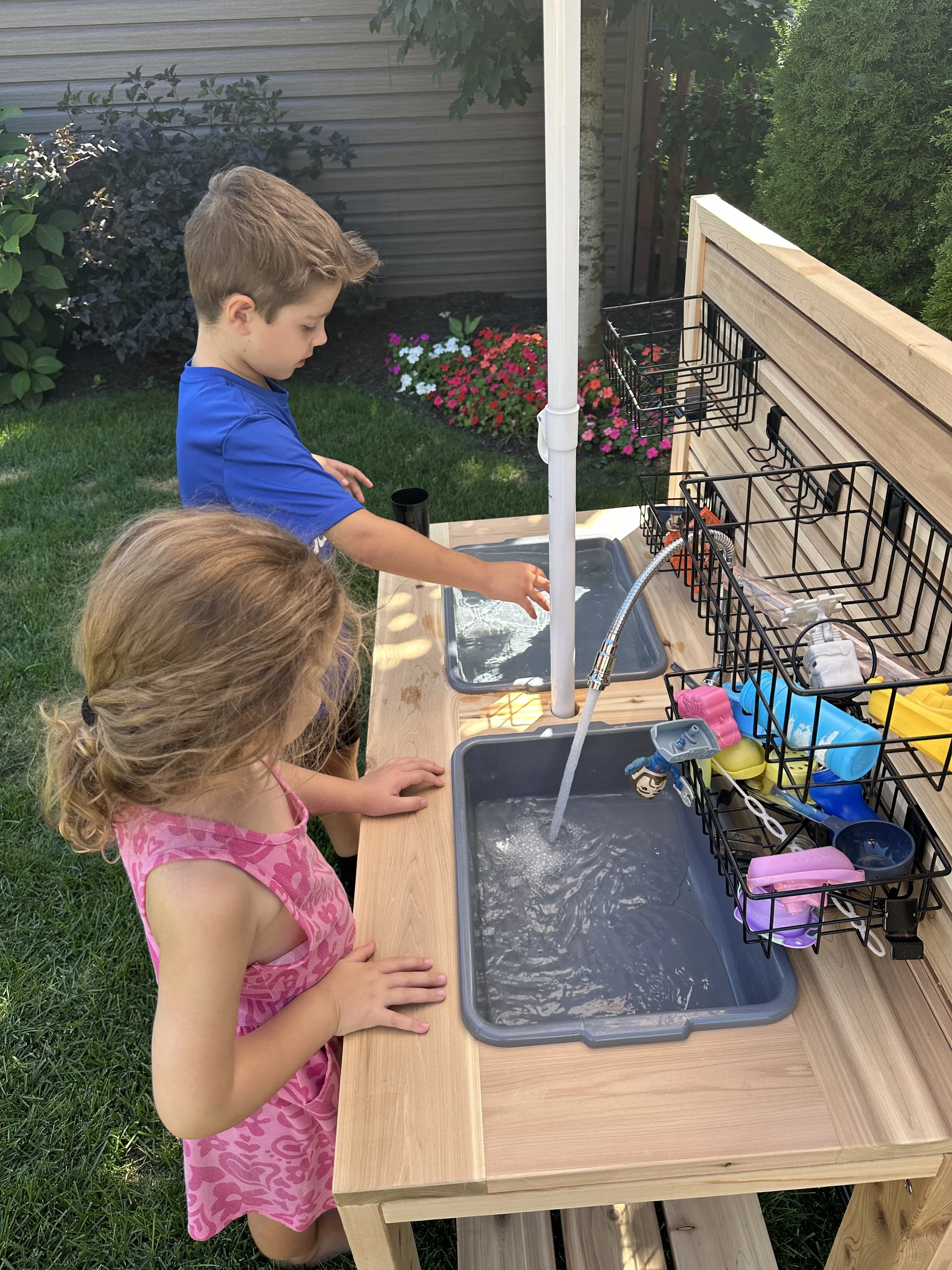Two children, a boy and a girl, are playing at an outdoor water play station with sinks and toy dishes.