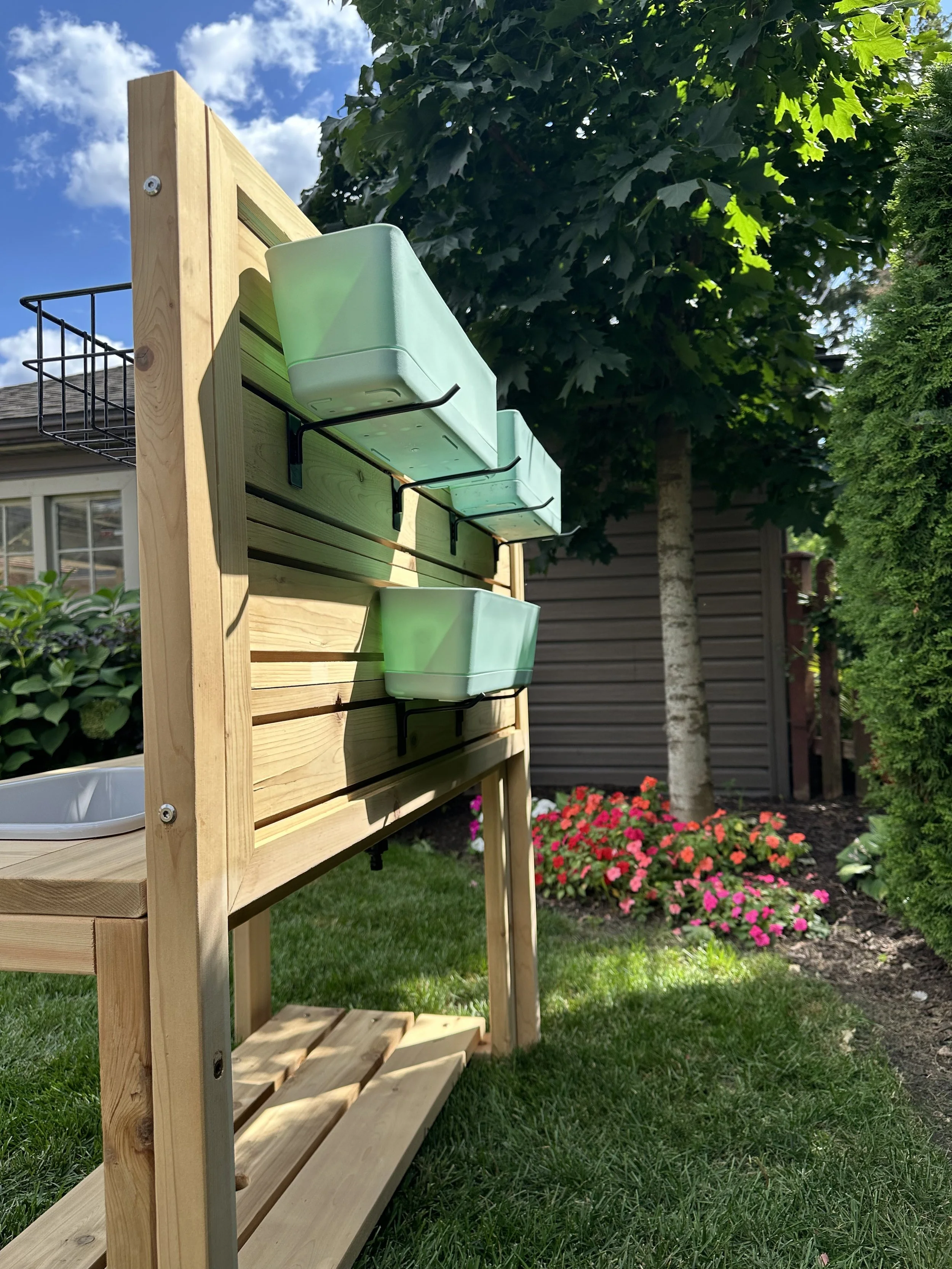A handcrafted Red Cedar sensory table featuring an integrated activity slat wall with hanging hooks and flower pots for a garden-themed play station.