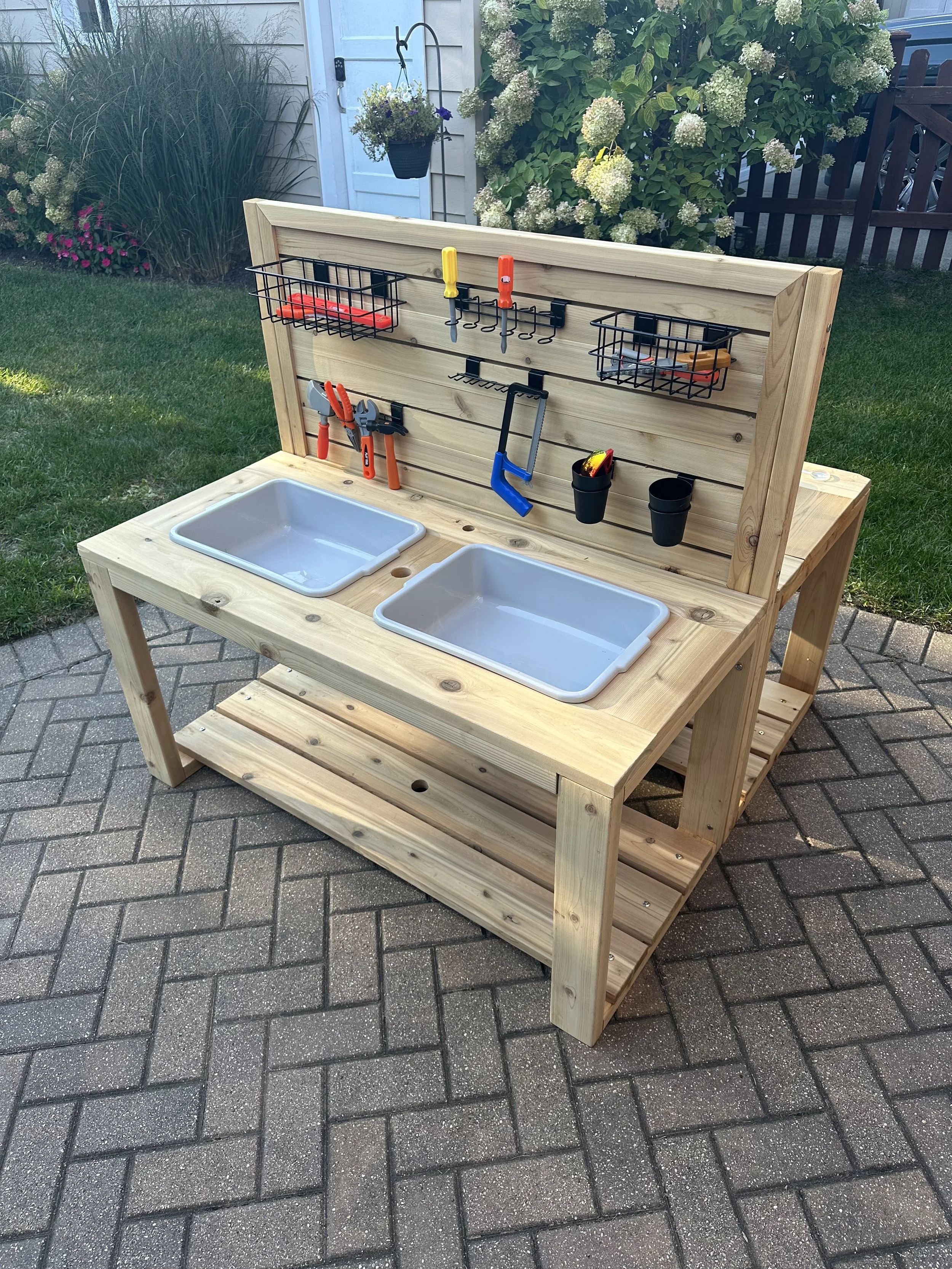 A wooden outdoor workbench with a pegboard back, featuring storage baskets, hooks, and tools. It has two white plastic sinks and is set on a brick patio in a garden with green grass, plants, and flowering bushes.