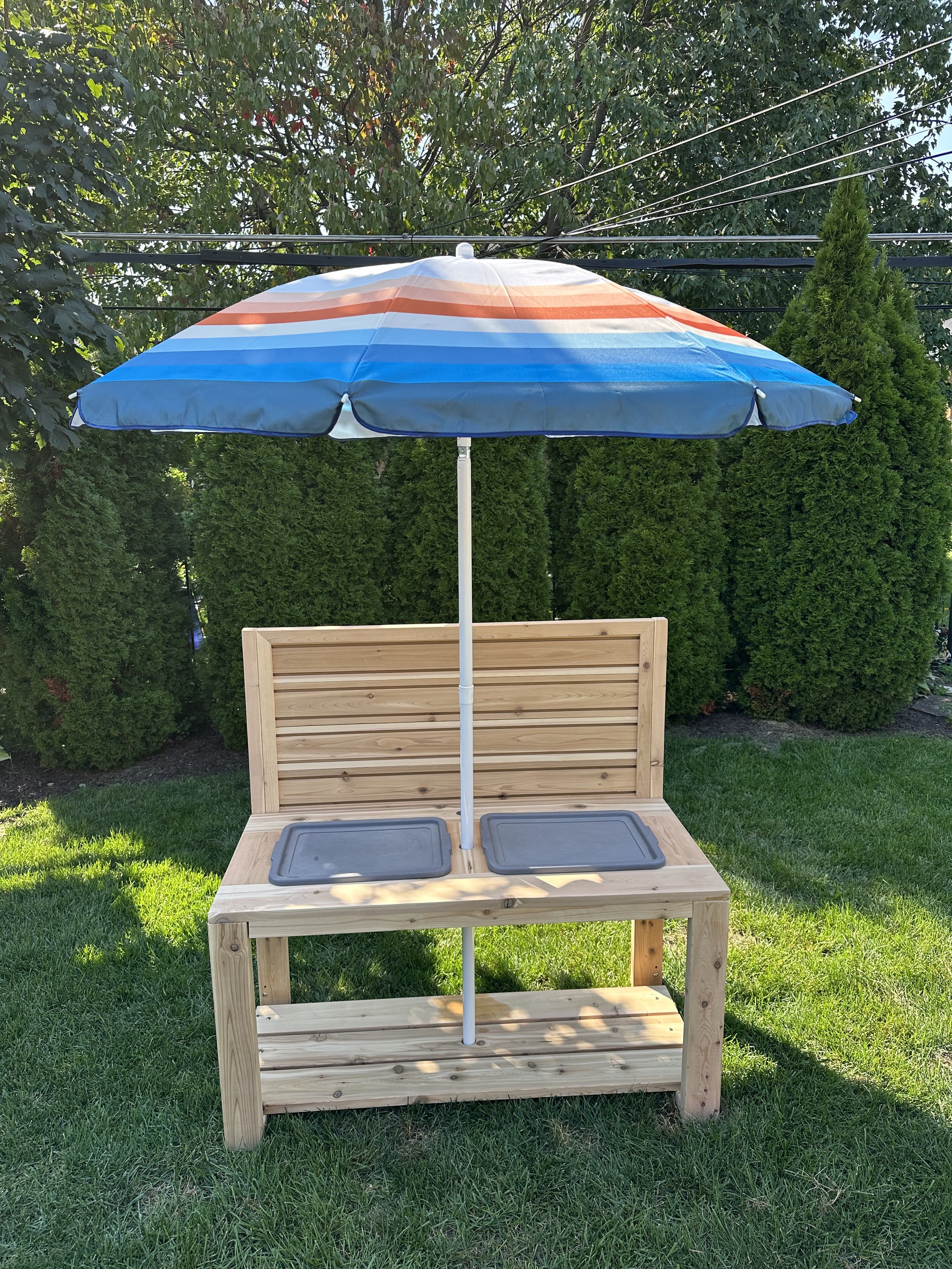 A wooden outdoor setup with a bench, two trays, and a large striped umbrella providing shade, situated on a grassy area with green bushes and trees in the background.
