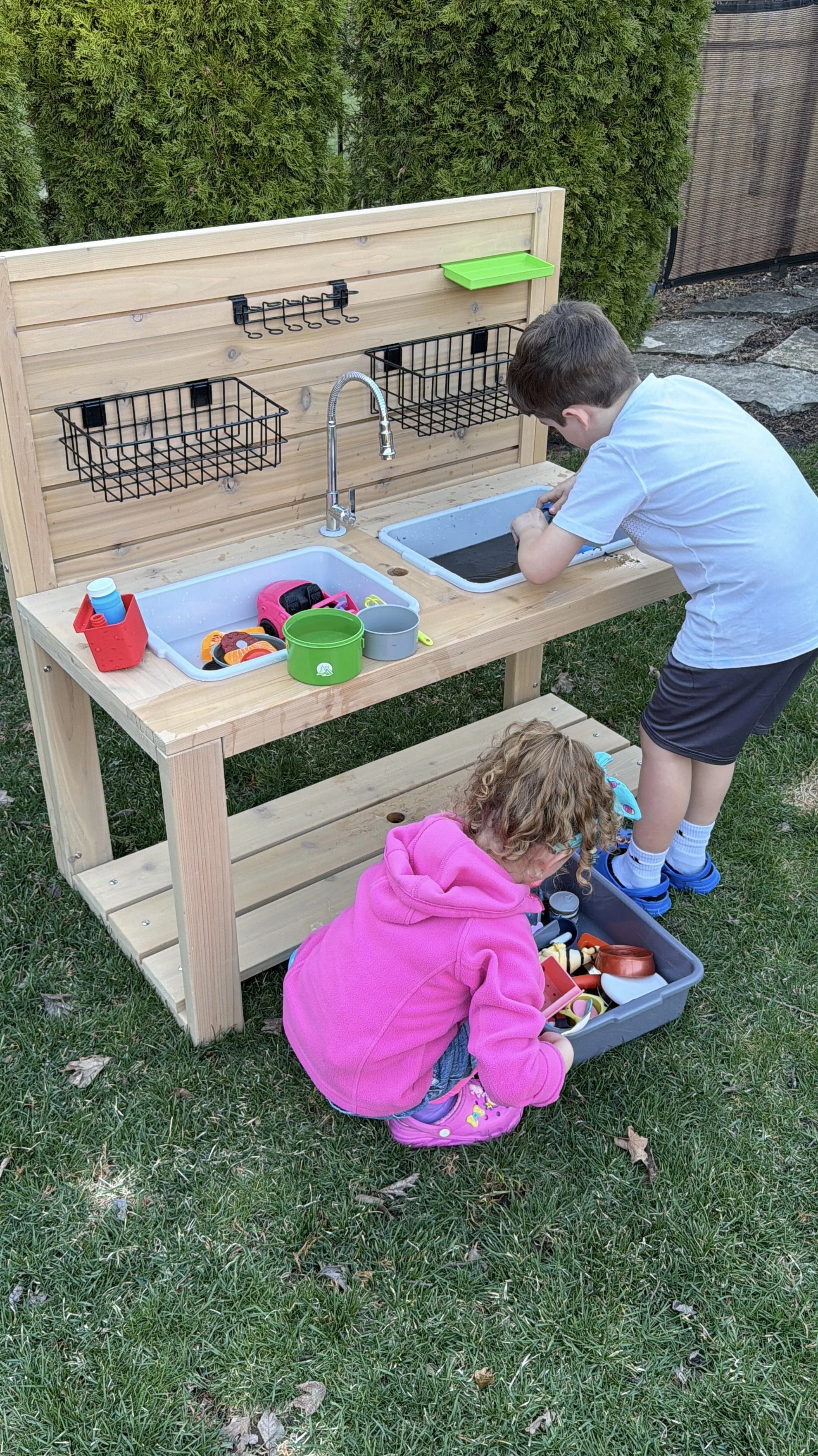 Two children playing at an outdoor play kitchen set with sinks, dishes, and utensils.