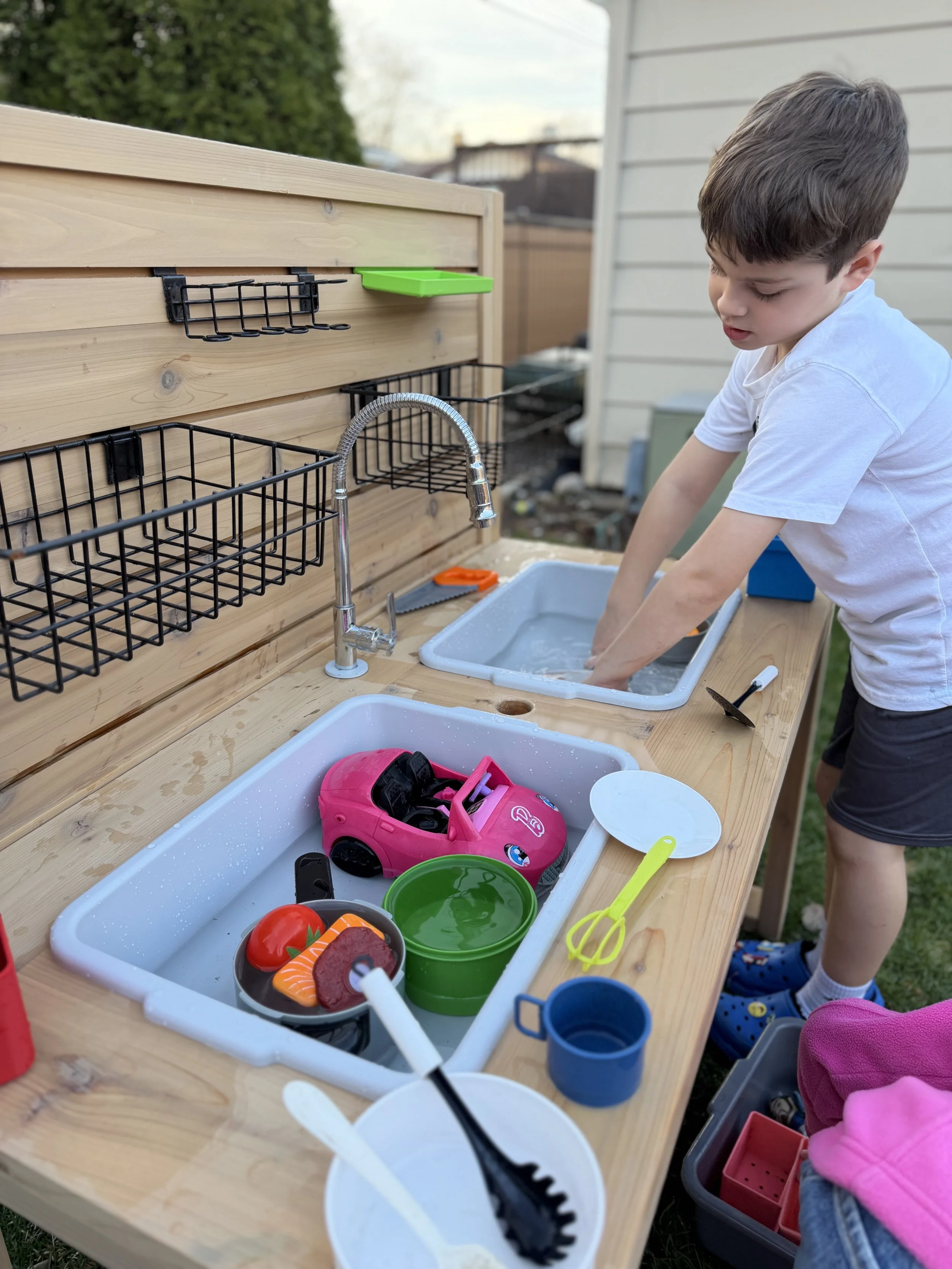 A Kinderbuilt cedar sensory table featuring a vertical activity slat wall with storage bins and a working water faucet.