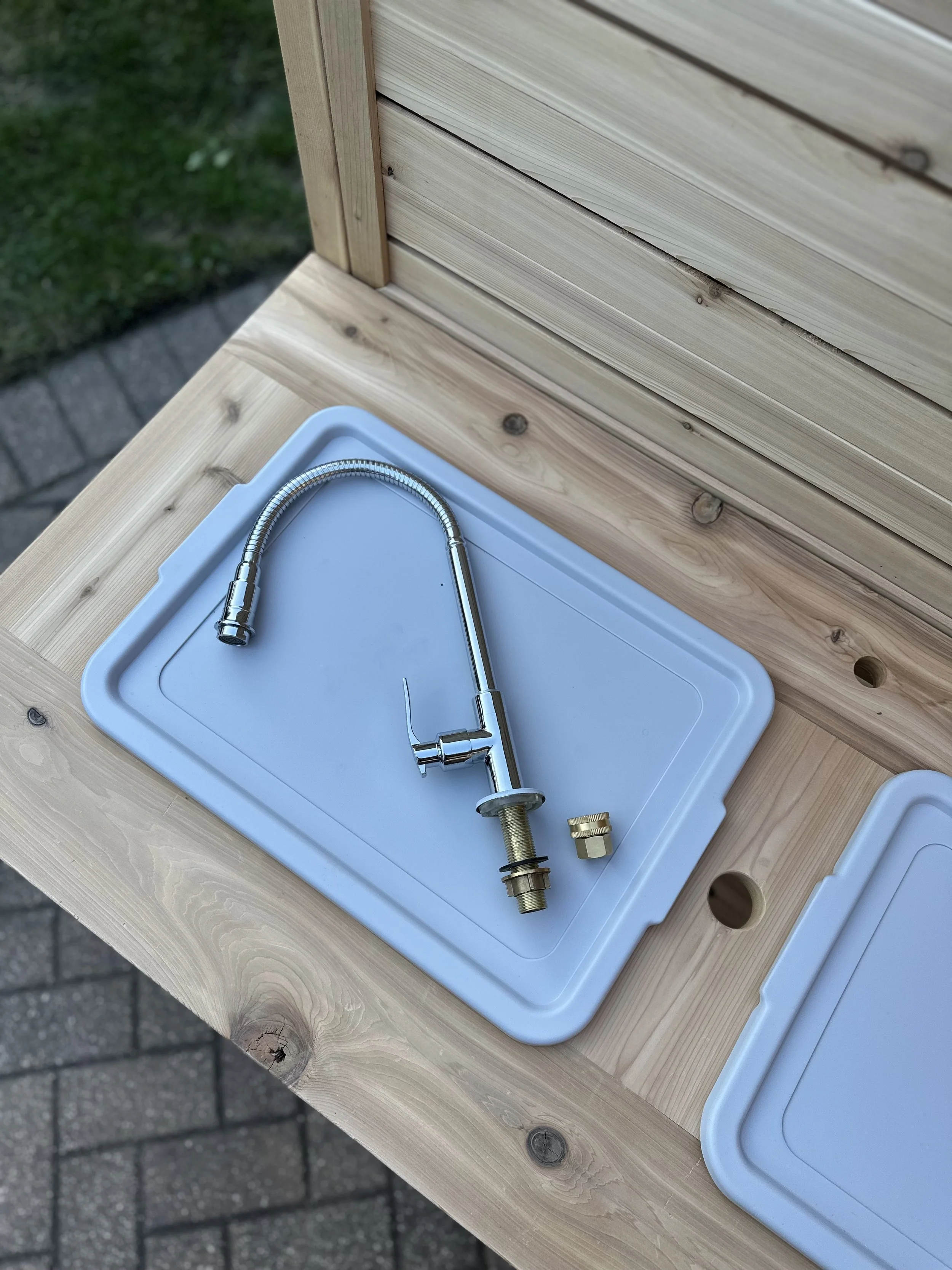 A stainless steel kitchen faucet with a flexible spring spout, a separate brass mounting piece, and a white plastic sink cover on a wooden worktable outside.
