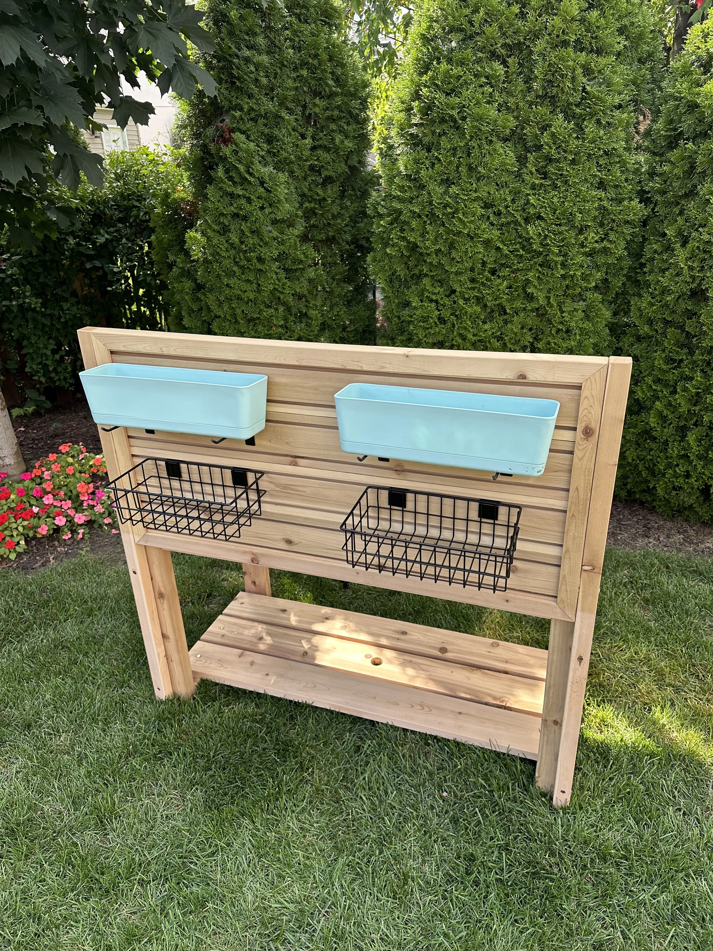 A wooden outdoor storage and planting stand with two light blue rectangular planters at the top, two black wire baskets underneath, and a lower shelf, set on a grassy yard with green bushes and flowers in the background.