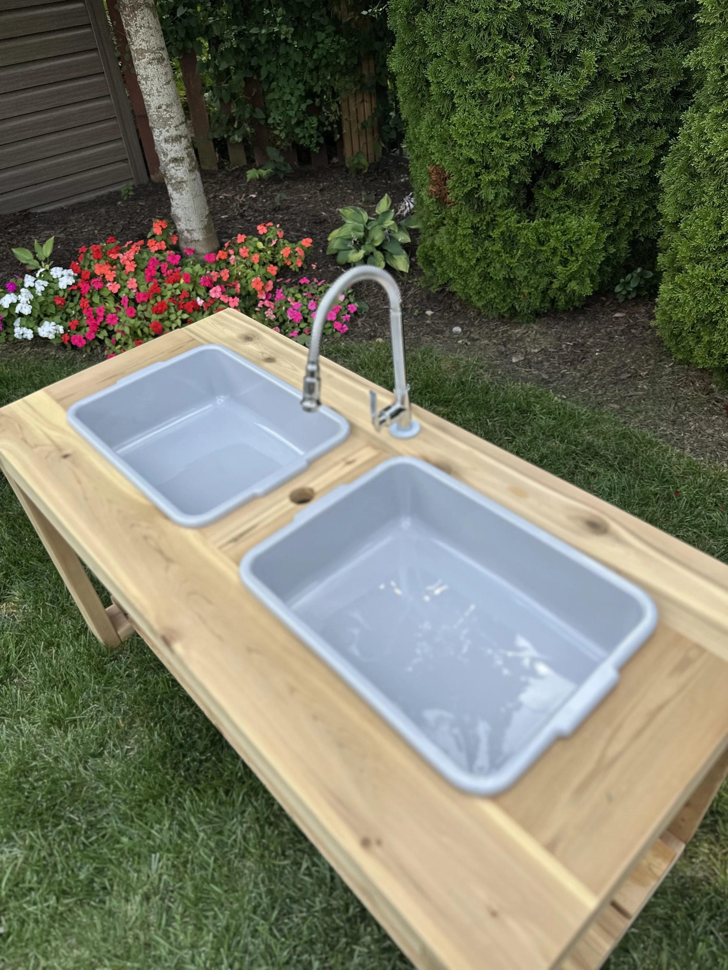 Outdoor wooden sink station with two plastic basins and a metal faucet, set in a garden with colorful flowers and green shrubbery.