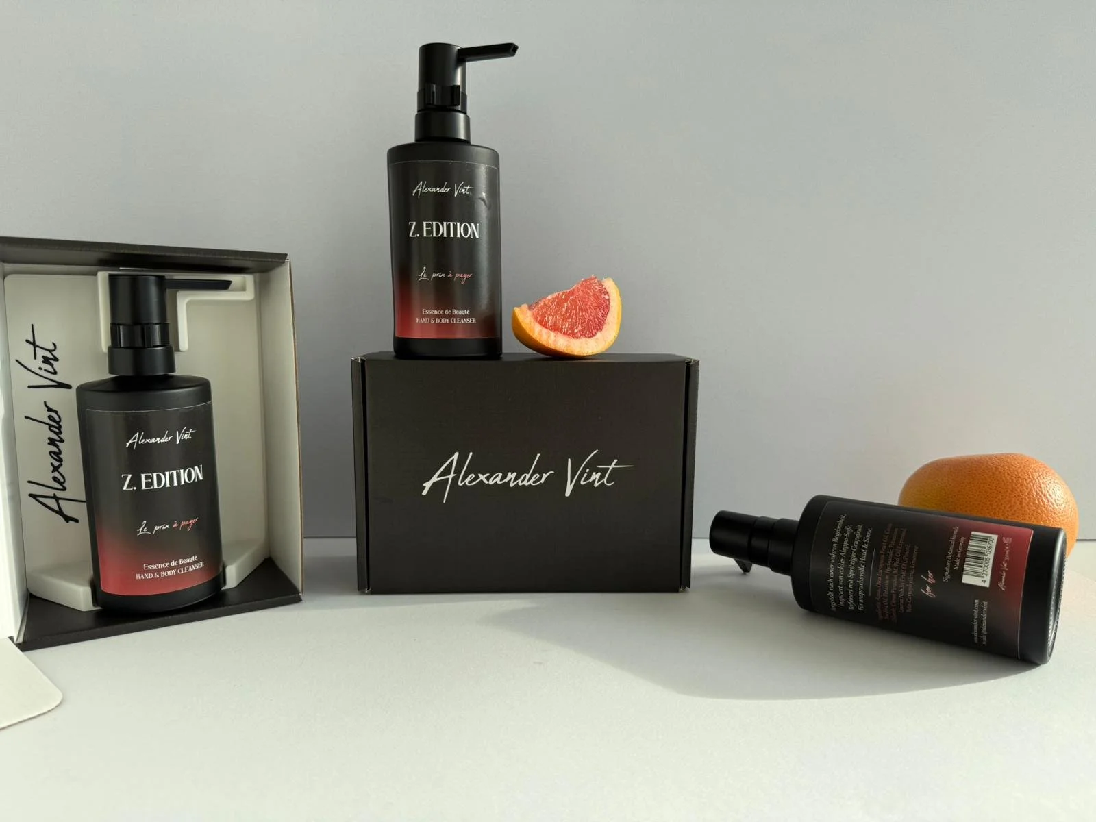 Drei Schwarze Flaschen mit Alexander Vint Z. EDITION Beschriftung, eine in einer weißen Box, eine auf einem schwarzen Kasten mit Alexander Vint Schrift, die dritte liegt schräg auf der Oberfläche, neben einer Grapefruit-Hälfte und einer ganzen Grapefruit.