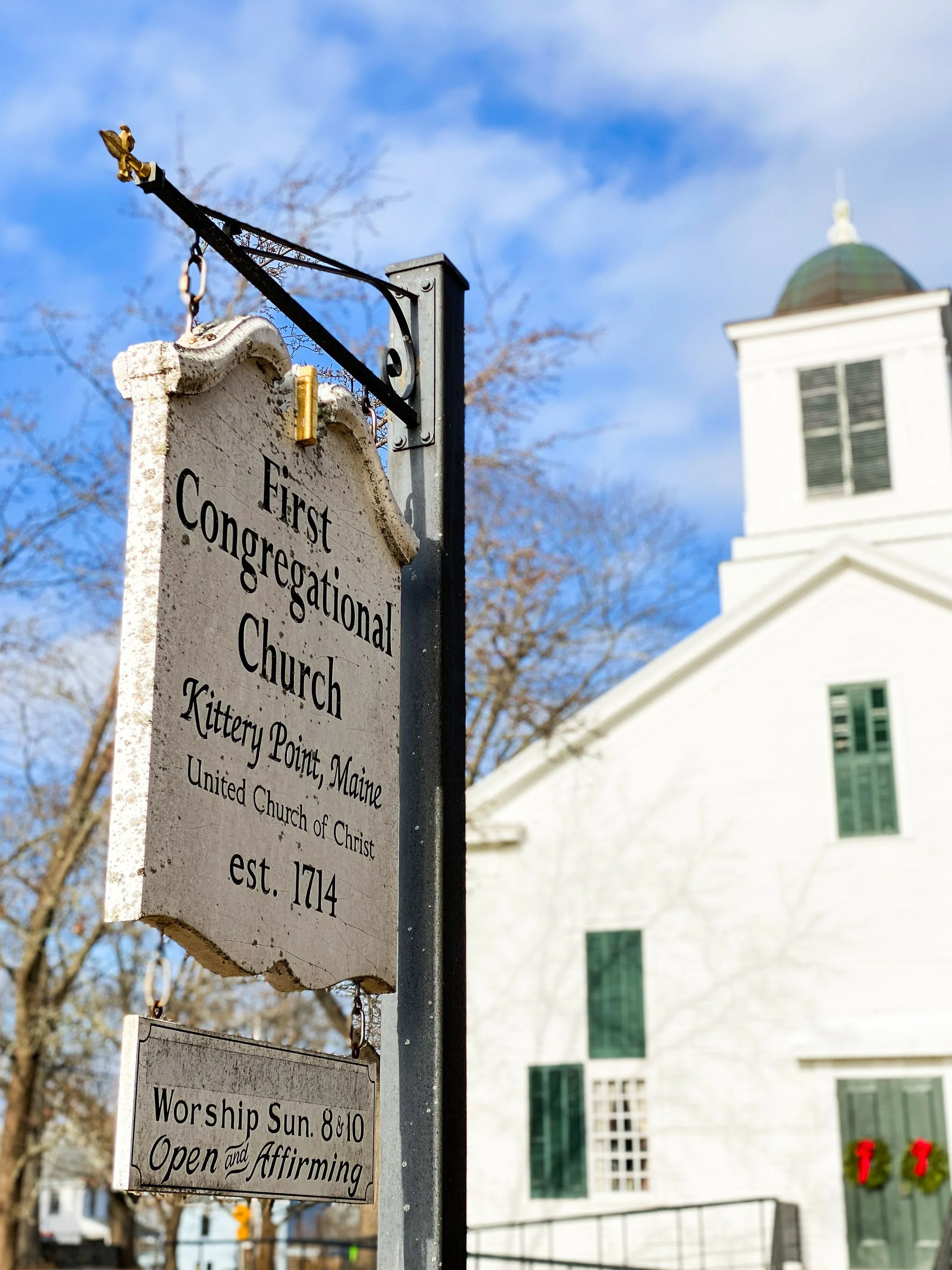 Historical church sign for the First Congregational Church in Kittery Point, Maine, established in 1714, with a background of the church building and blue sky.