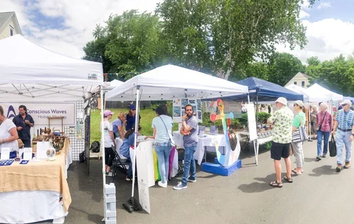 People browsing outdoor market stalls with tents, trees in background, and vendors selling various goods.