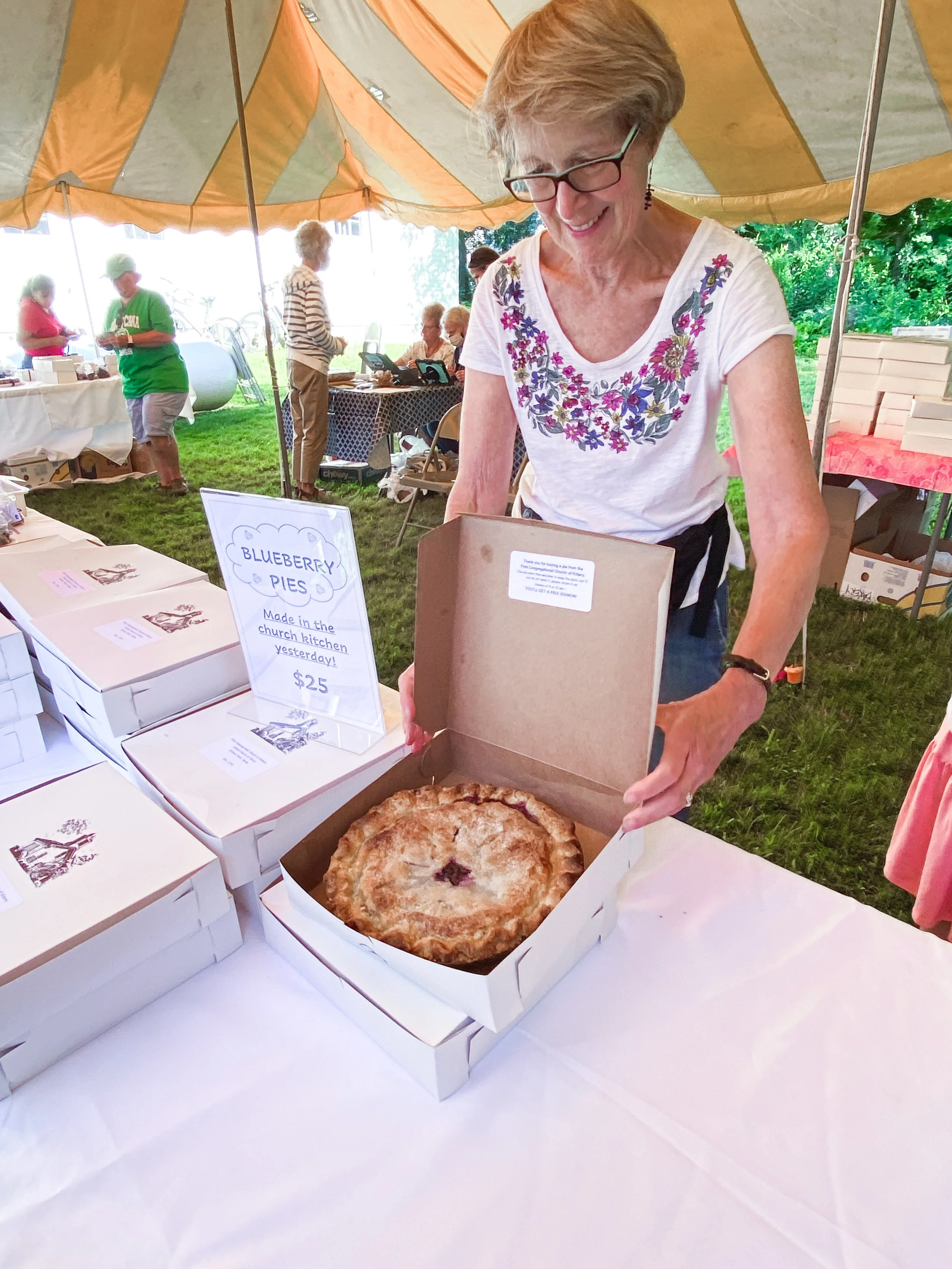 A woman with short gray hair, glasses, and a white shirt with floral embroidery, smiles as she opens a pizza box containing a blueberry pie on a white table. Behind her, there are other people and tables under a large yellow and orange canopy at an outdoor event.