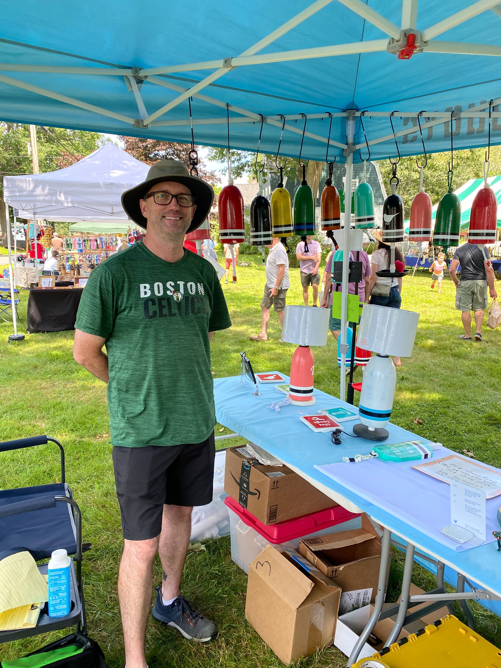 A smiling man wearing glasses, a green T-shirt, shorts, sneakers, and a wide-brimmed hat standing beside a booth with assorted colorful buoy floats hanging above. The booth is at an outdoor market with other stalls and people browsing in the background, under a blue canopy.