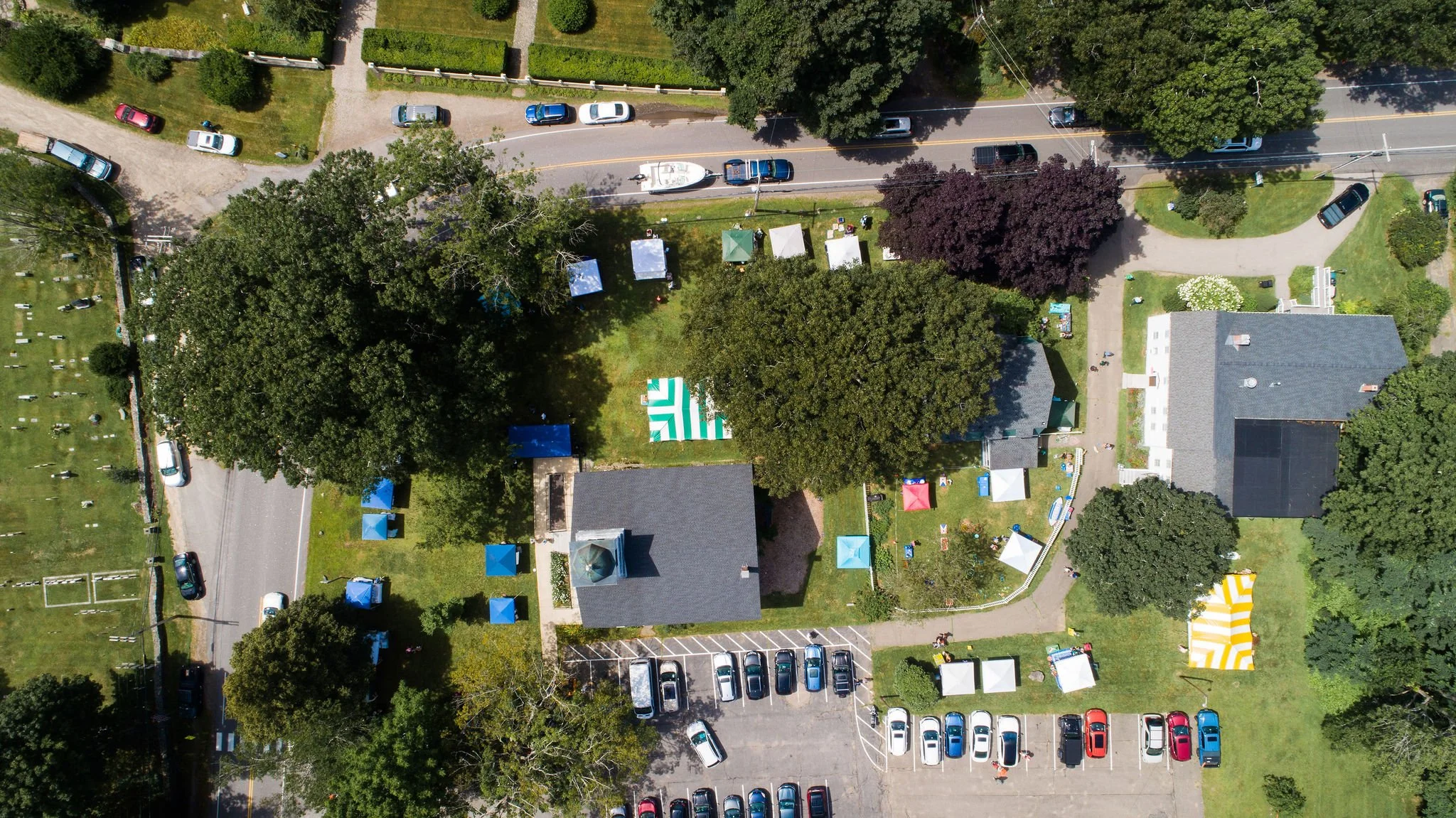 An aerial view of a neighborhood with houses, trees, and parked cars, along with tents and outdoor setups in a backyard, indicating a gathering or event.