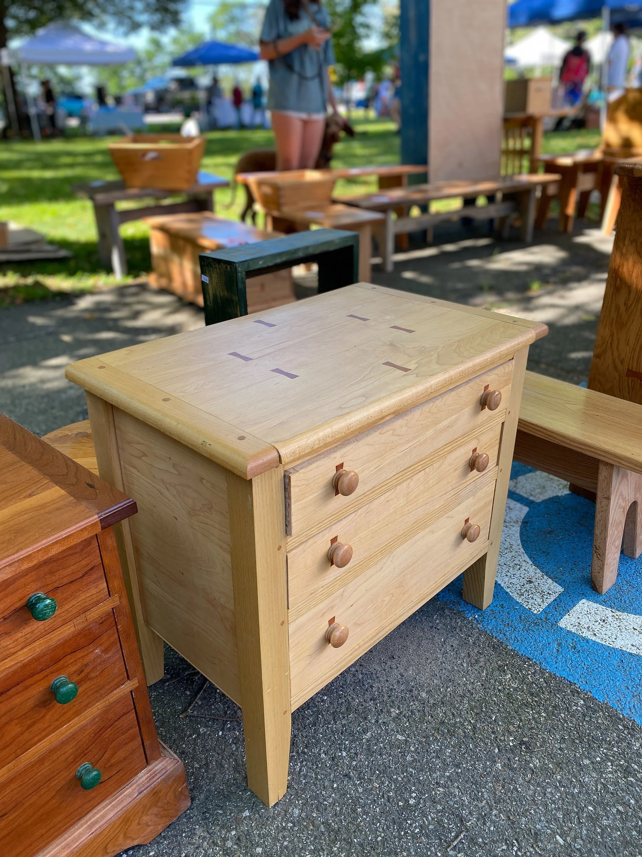 A wooden dresser with three drawers and round knobs, placed on pavement at an outdoor market with other furniture and people in the background.