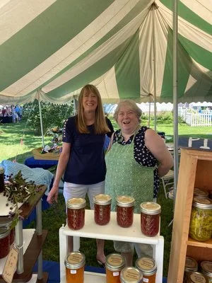 Two women smiling at a craft stall at an outdoor fair, with jars of homemade jam and honey displayed on the table, under a large green and white striped tent. Blueberry Festival First Congregational Church Kittery Point
