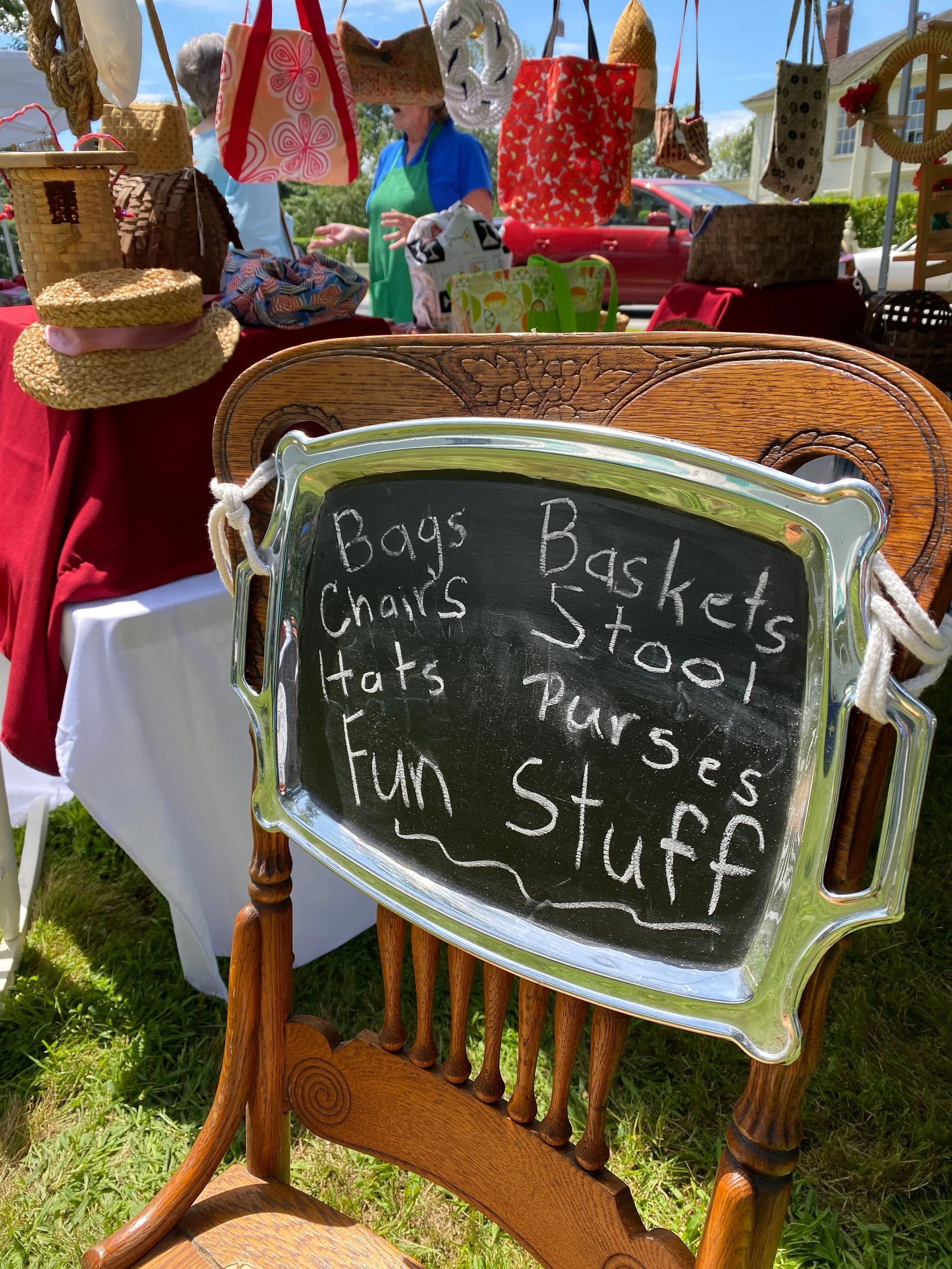 A wooden chair with a chalkboard sign attached to its back. The sign has handwritten text listing various items for sale, including bags, chairs, hats, and fun stuff. In the background, there are tables with assorted bags, hats, and other items displayed at an outdoor market or yard sale. There are people browsing and a house visible in the distance.