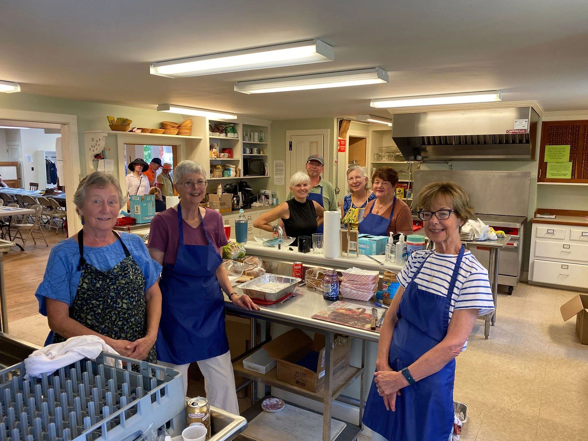 Group of seniors working in a community kitchen, smiling and wearing aprons, with shelves of kitchen supplies in the background.