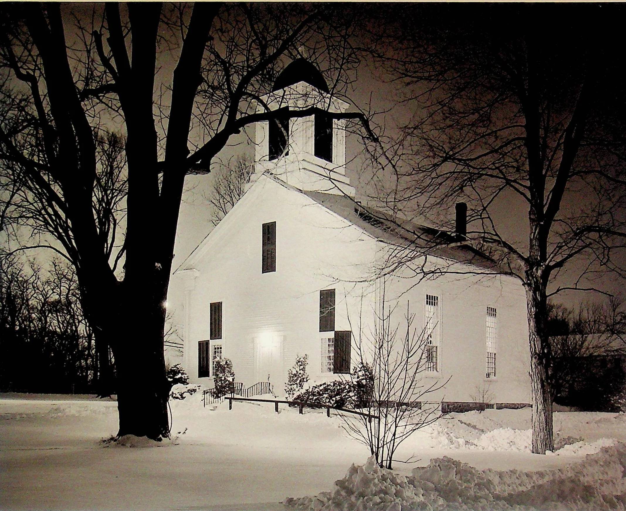 A white church building surrounded by snow with tall, leafless trees around it, illuminated at night.