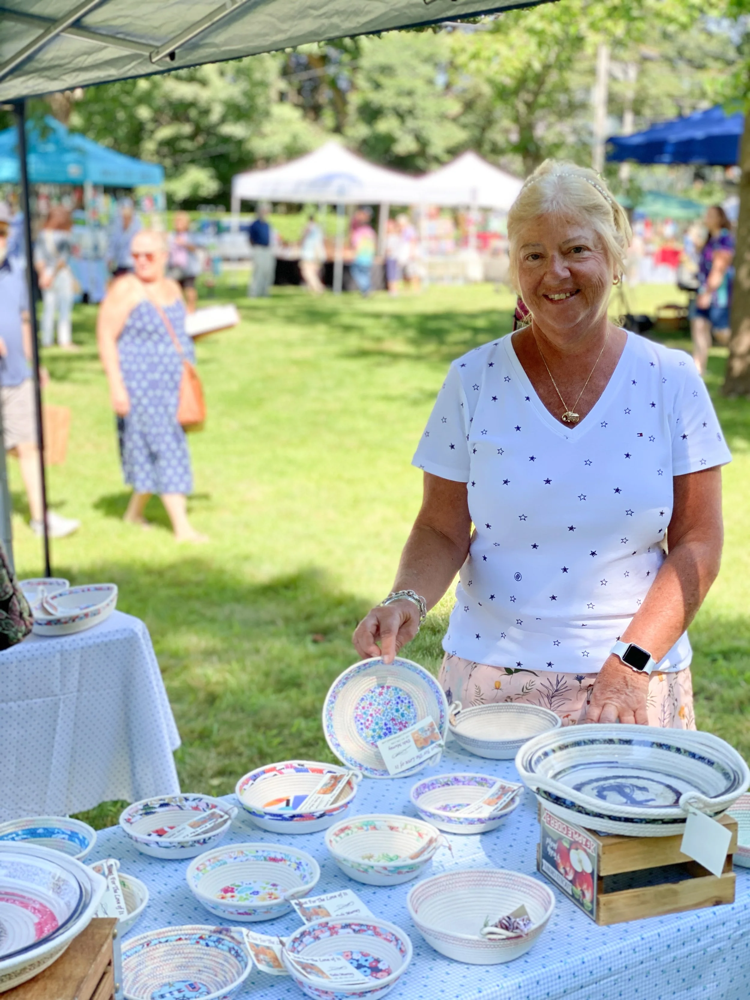 A woman standing at a table displaying decorated pottery bowls at an outdoor market or craft fair with tents and people in the background.