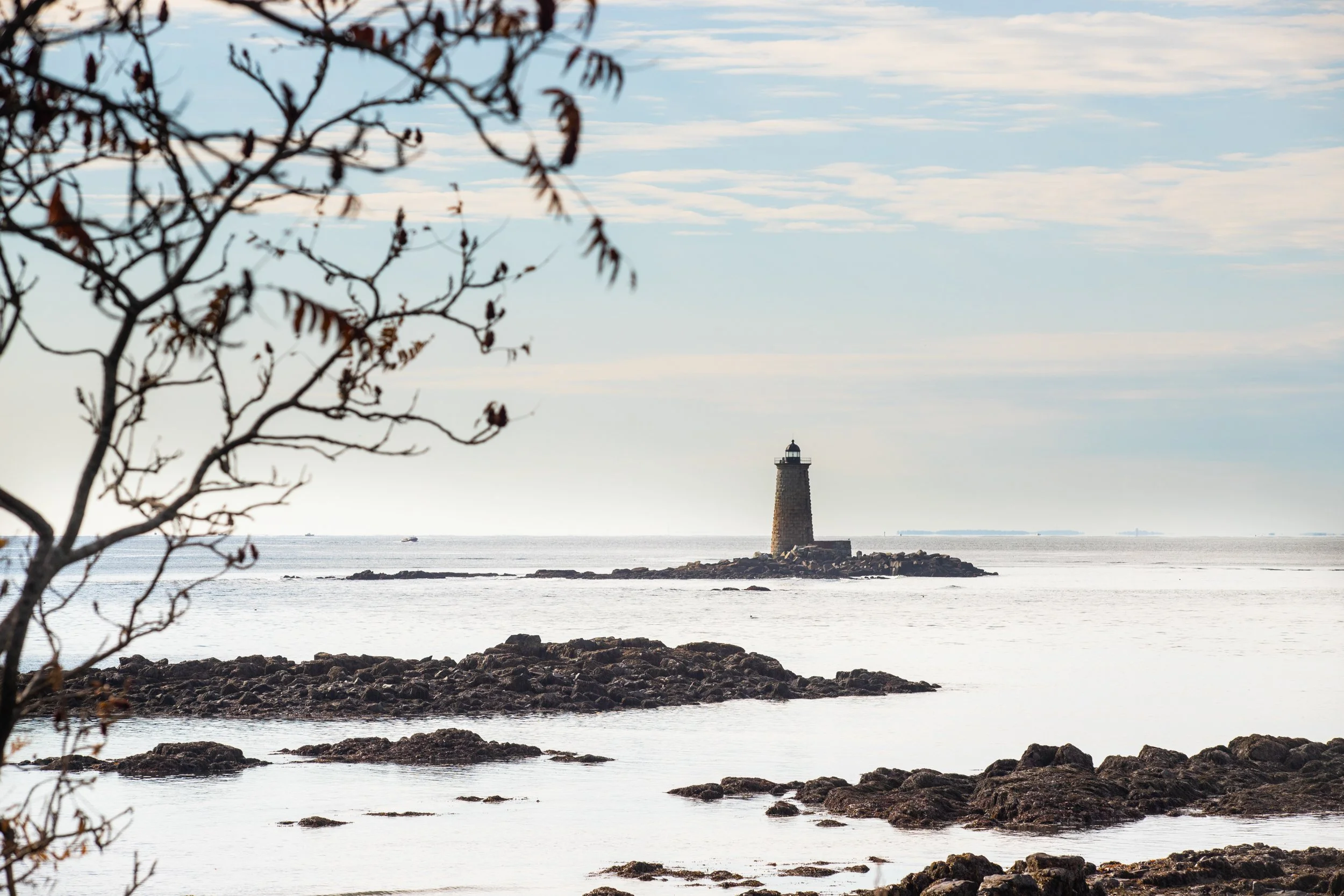A lighthouse on a small rocky island off the coast, with water in the foreground, a few scattered rocks along the shore, and a sky with scattered clouds.