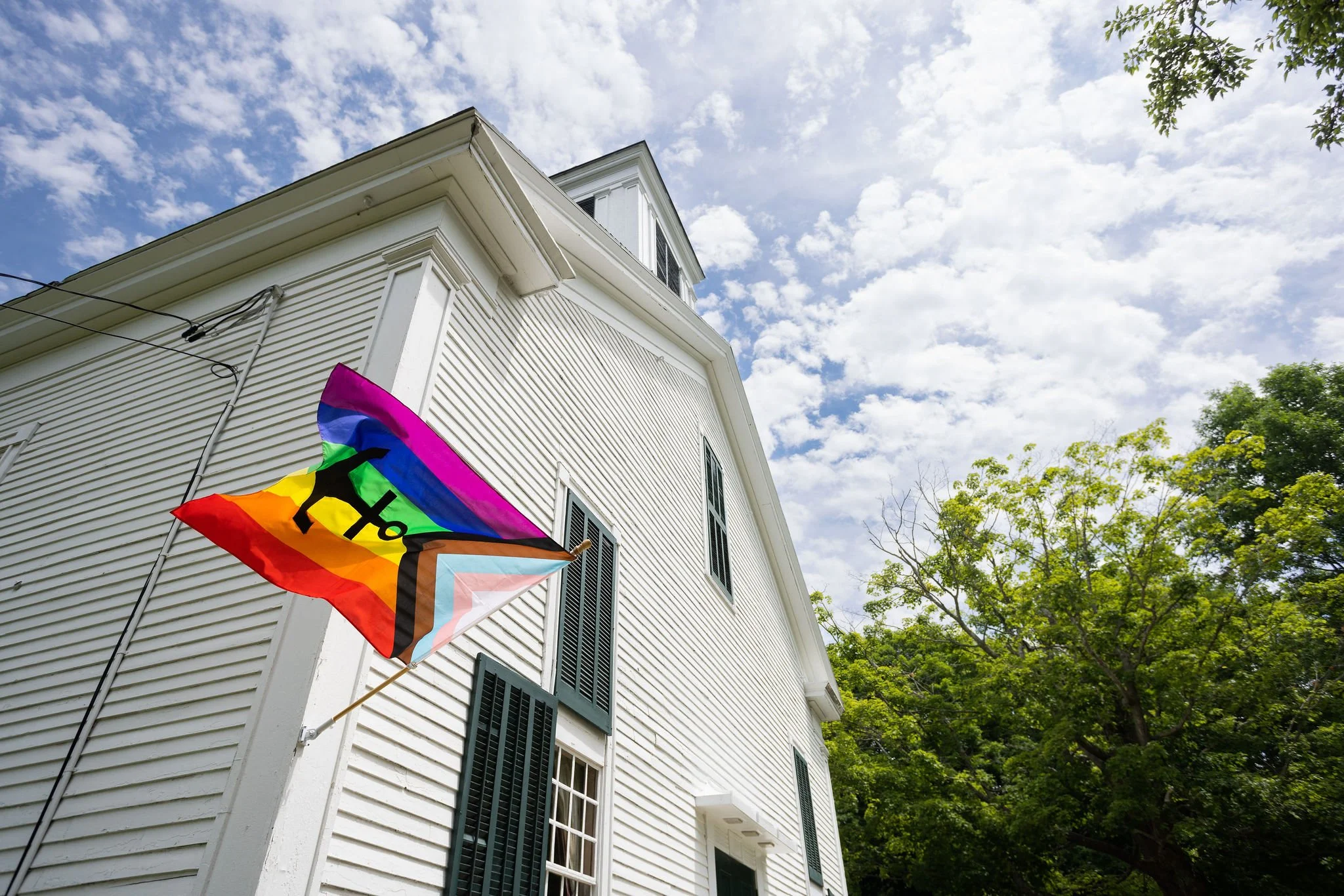 White house with black window shutters, a rainbow and black pride flag flying outside, and trees with green leaves, under a partly cloudy sky.