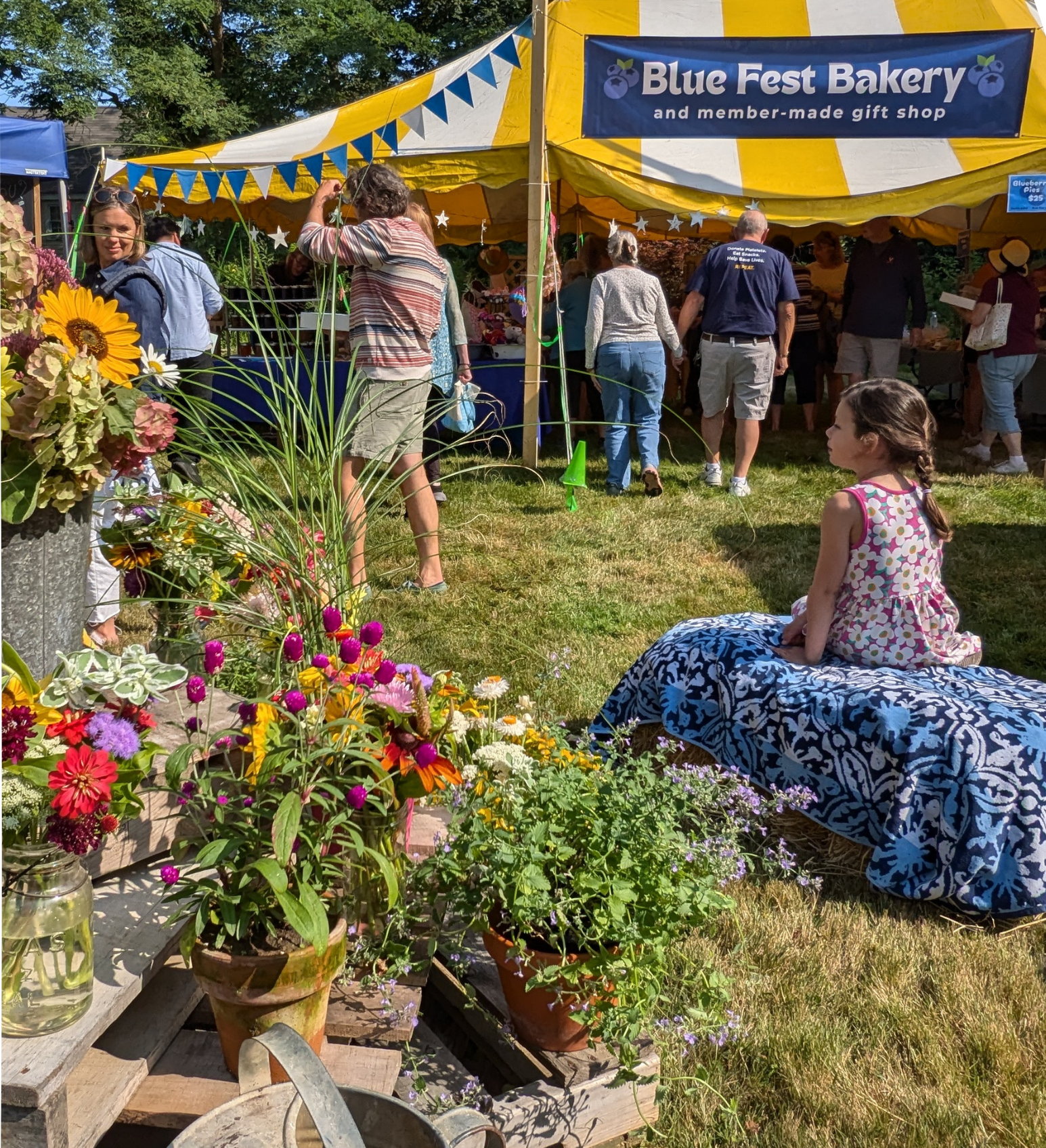 People visiting Blue Fest Bakery and gift shop at an outdoor event, with colorful flowers in the foreground and a yellow and white striped tent in the background. Blueberry Festival First Congregational Church Kittery Point