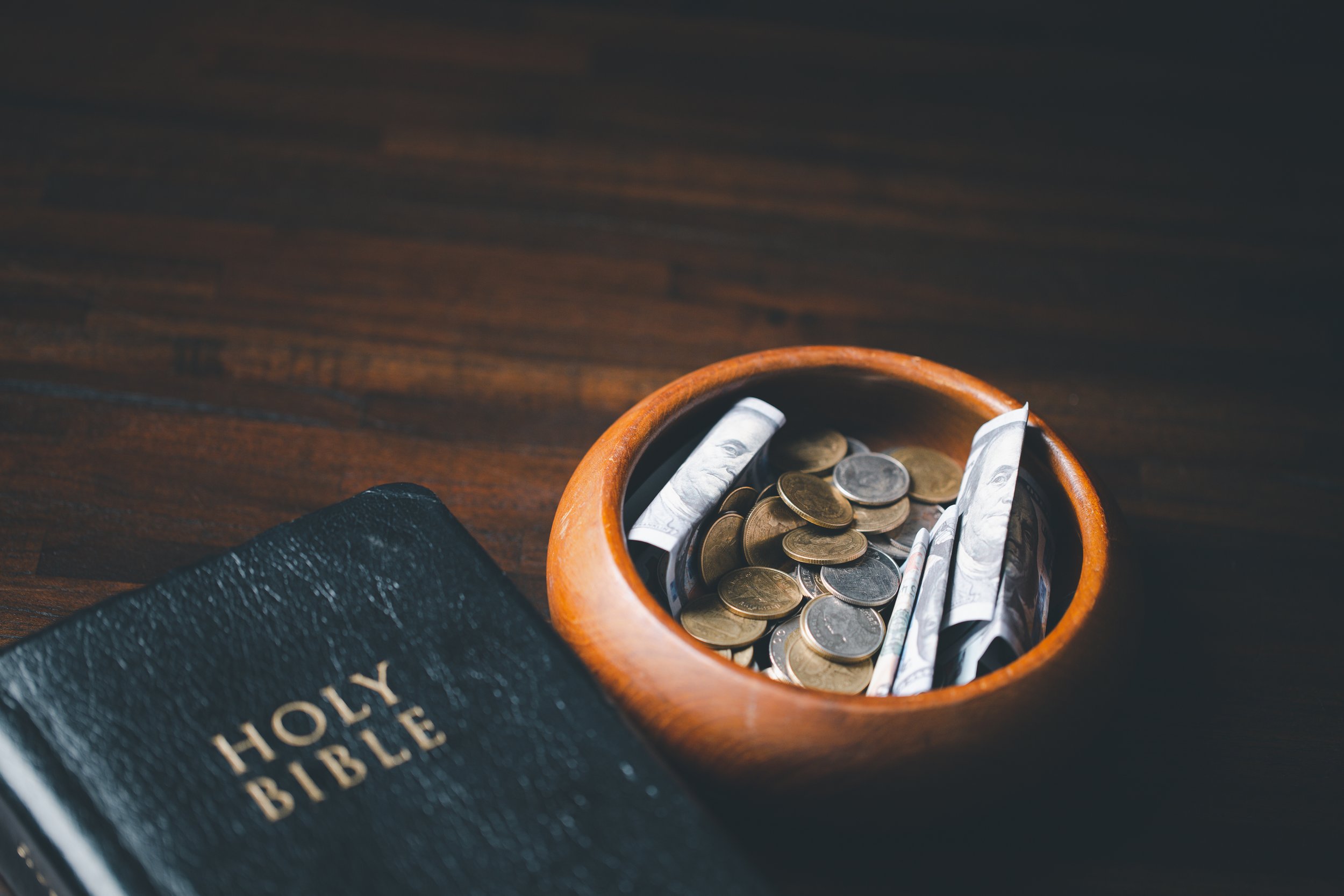 A black Holy Bible with gold lettering next to a small wooden bowl filled with coins and rolled dollar bills, placed on a dark wood surface.