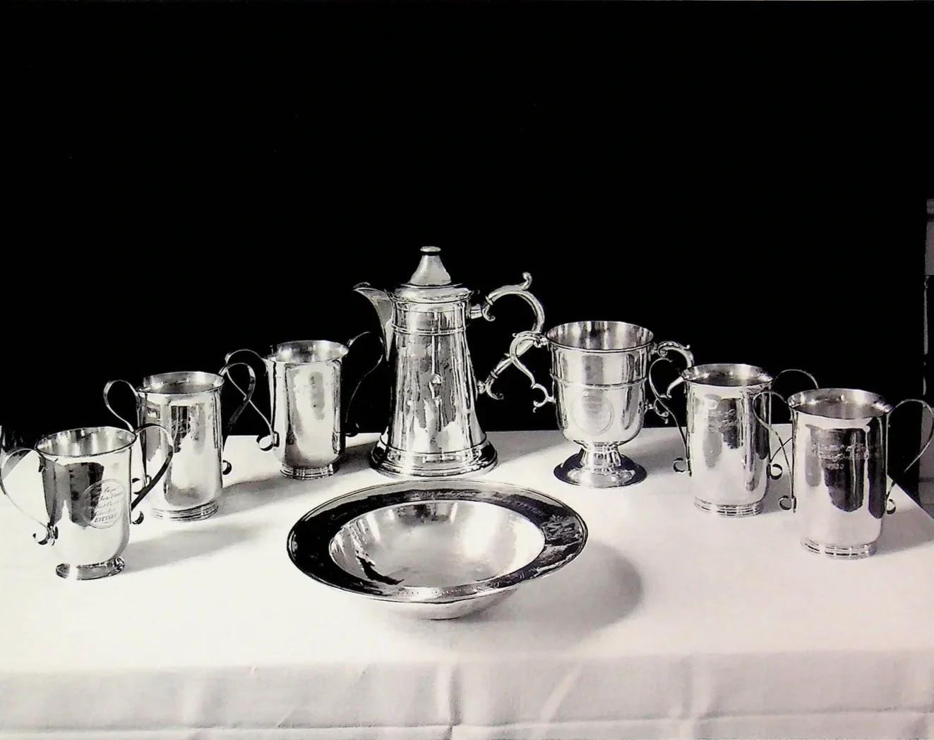 Set of silver tea and coffee serving pieces including a teapot, sugar bowl, creamer, cups, and a plate, arranged on a white tablecloth against a black background.