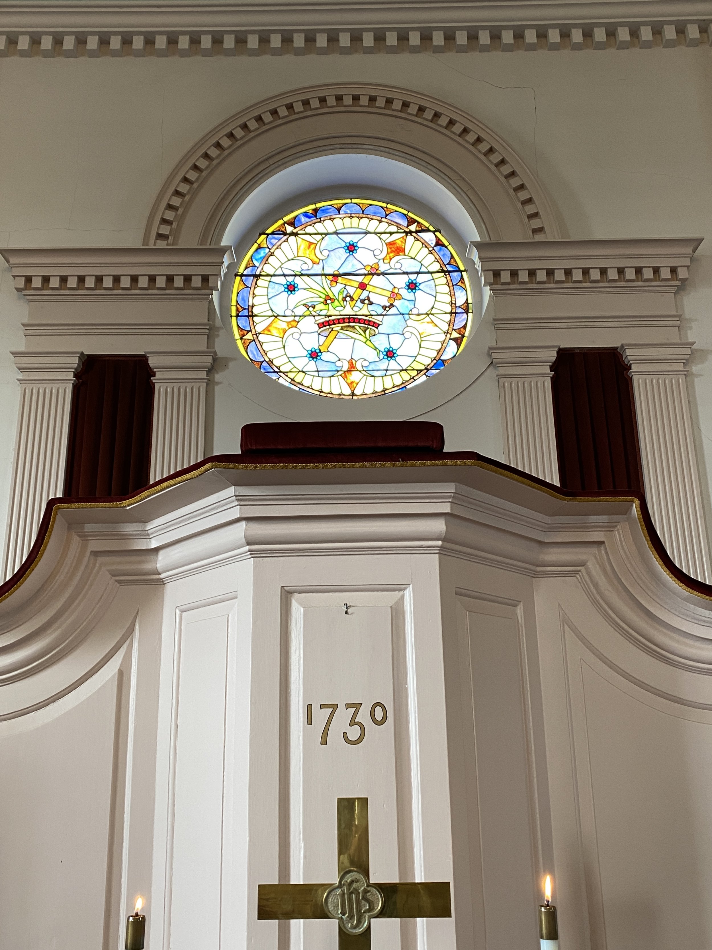 Inside view of a church pulpit with a stained glass clock above it showing the time as 11:22 and a white wall with decorative molding.