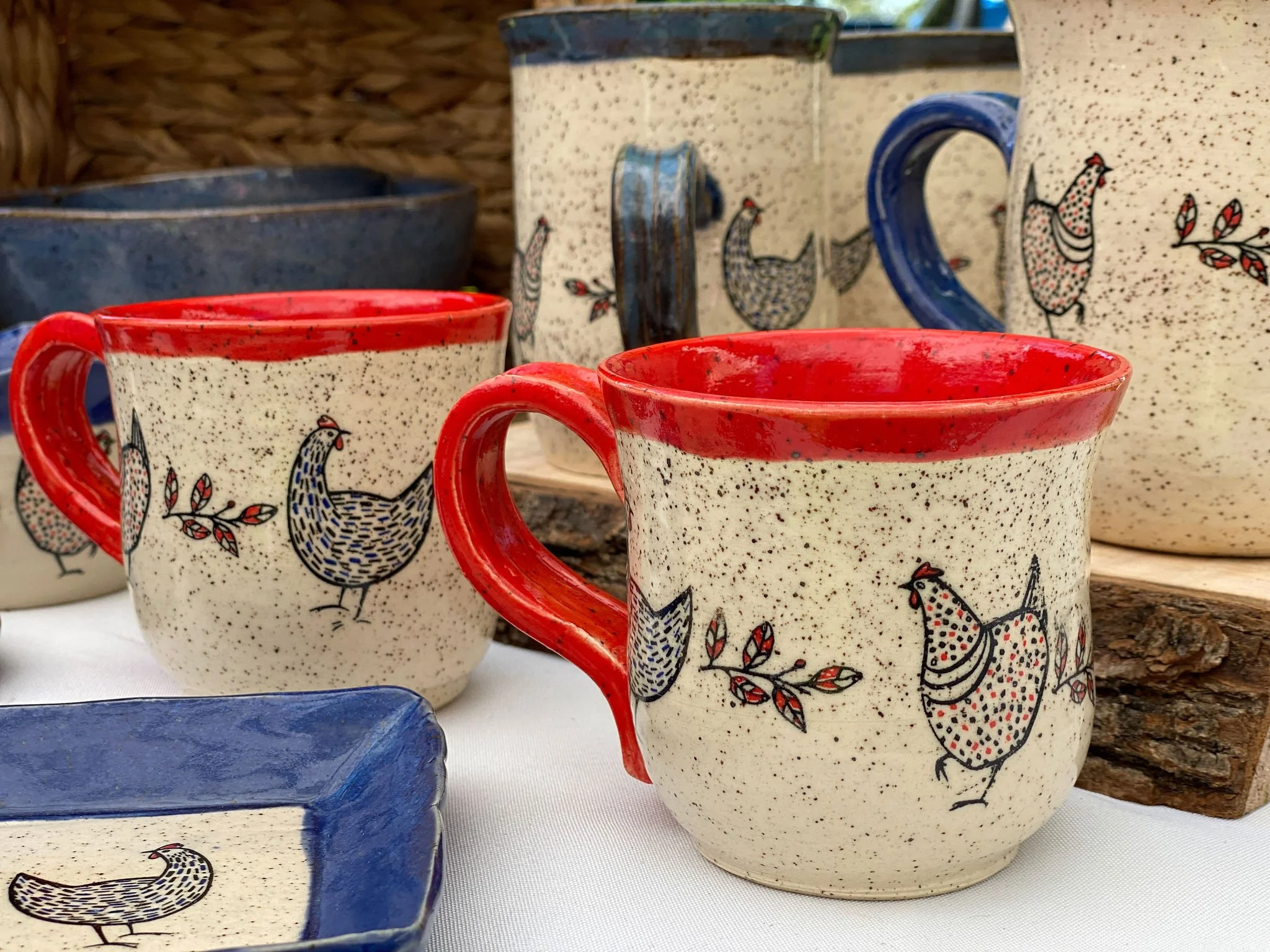 Set of ceramic mugs and bowls with a rooster and leaf pattern, mainly in white with black dots, and red, blue, and black accents.