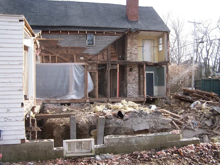 House undergoing demolition or renovation with exposed wooden framework and construction debris in the yard.