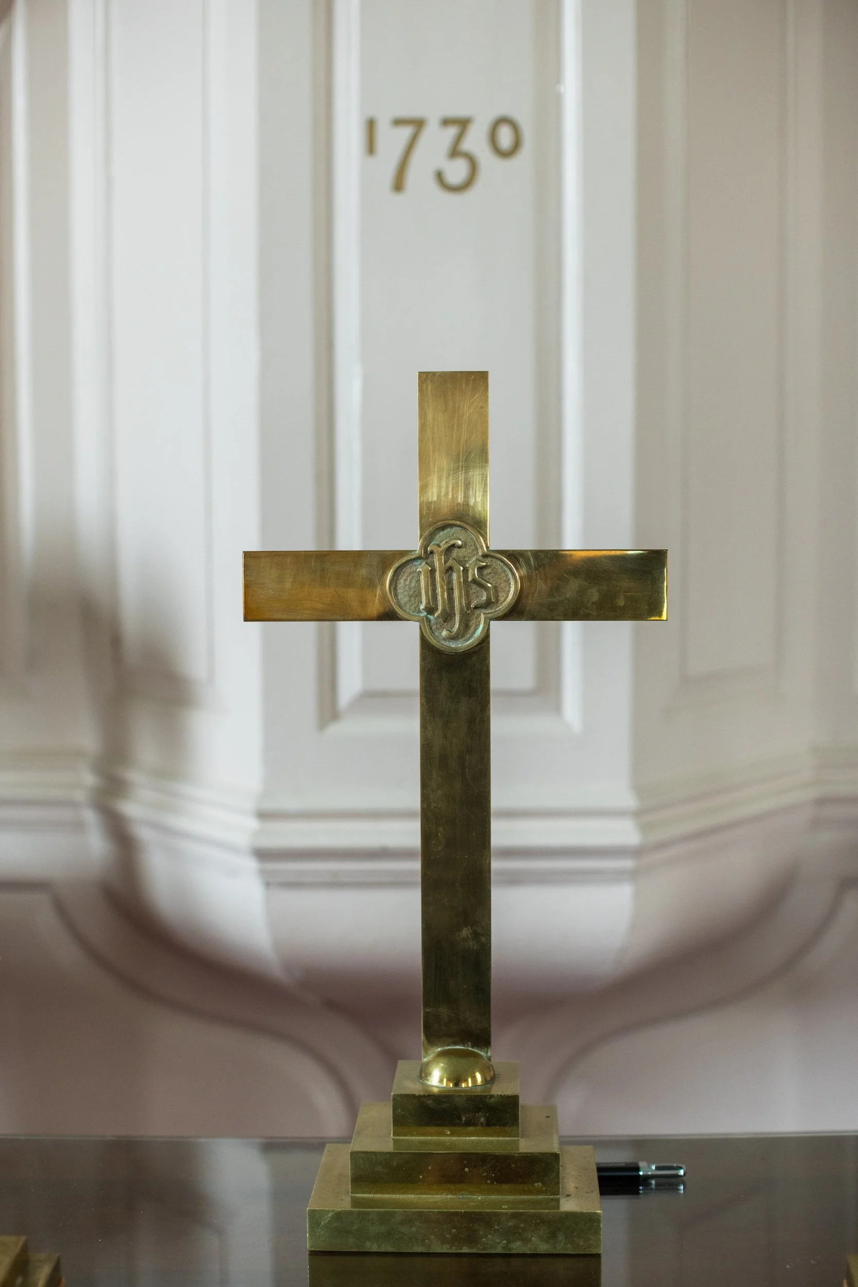 A brass cross with the letters IHS in a shield design at the center, placed on a table, with a white door and the number 1730 in the background.