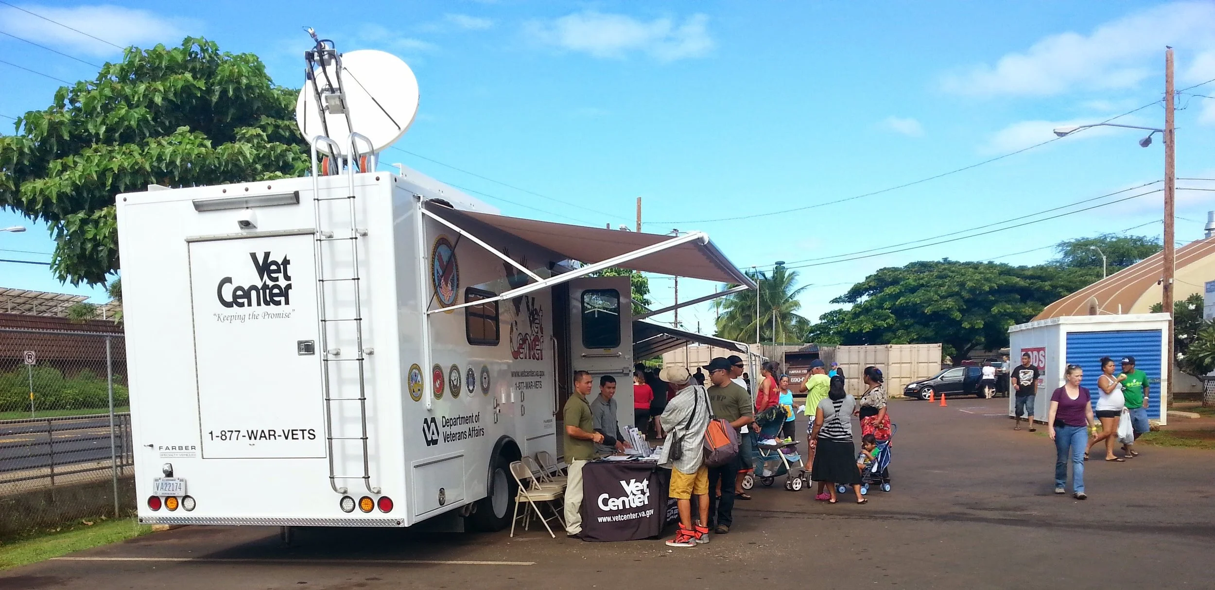 A mobile Vet Center truck parked outdoors with people gathered around it, some with strollers, at a community event on a sunny day with blue skies and trees in the background.