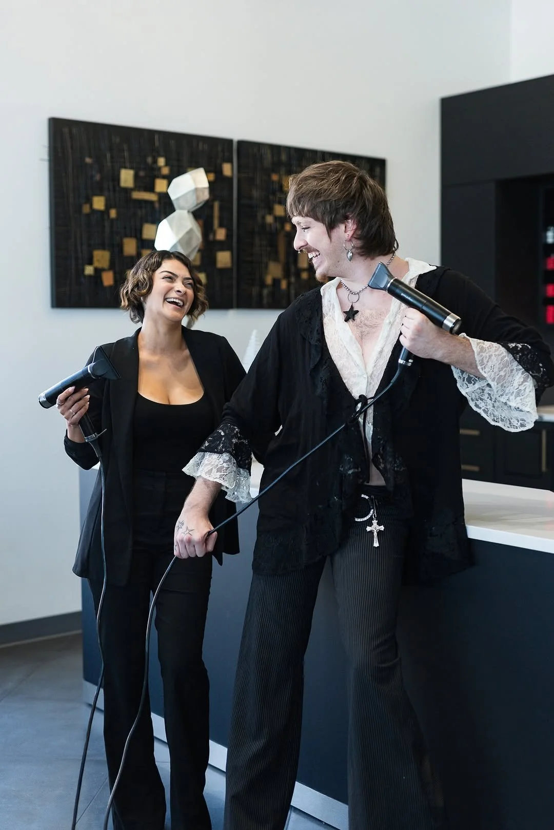 Two women sharing a laugh while holding hair dryers in a modern room with artwork on the wall.