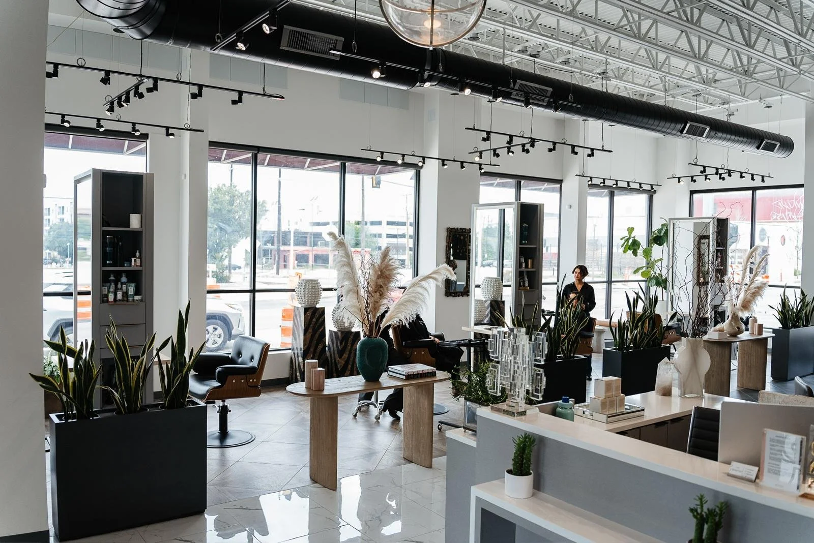 Interior of a modern hair salon with large windows, black and white decor, stylish furniture, and various plants.