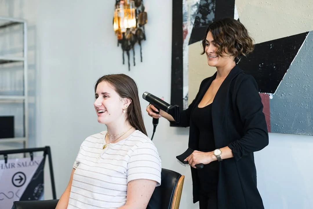 A hairstylist blow-drying a woman's hair at a salon.