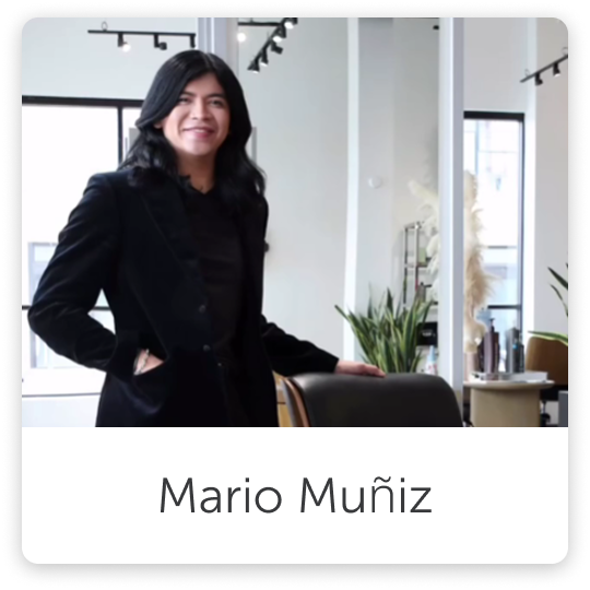 A man with long, dark hair wearing a black blazer and black top standing in a modern, well-lit room with large windows, indoor plants, and contemporary decor.