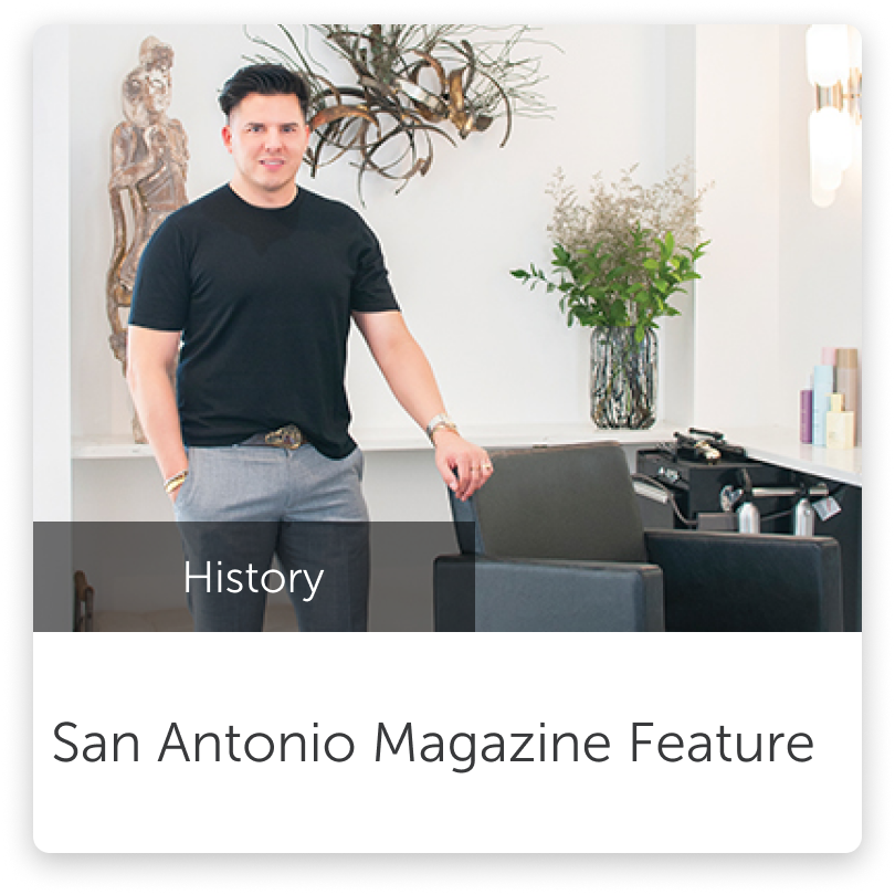A man in a black t-shirt and gray pants standing in a modern, stylish salon or studio. The background includes artwork, a large vase with greenery, and styling tools on a counter. The image appears to be part of a magazine feature on San Antonio.